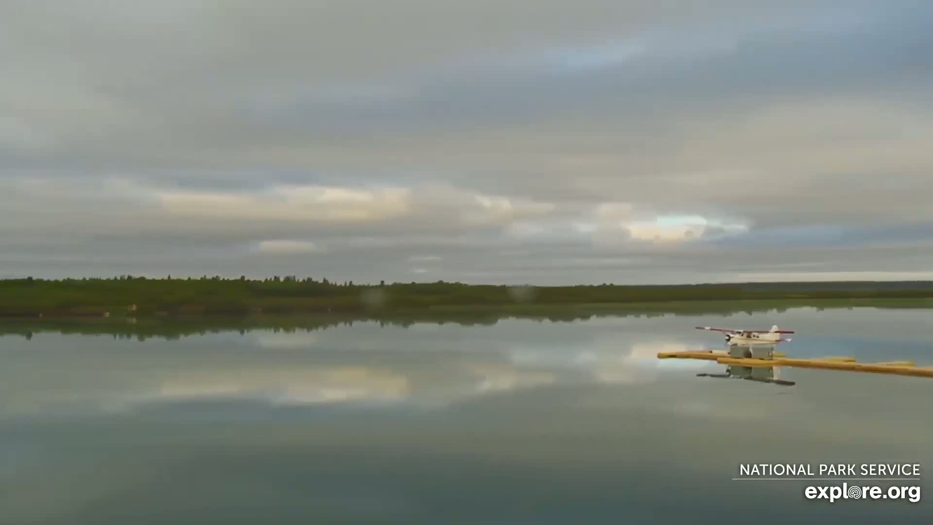 A seaplane sits on a calm lake under a mostly cloudy sky, with a treeline visible in the background.