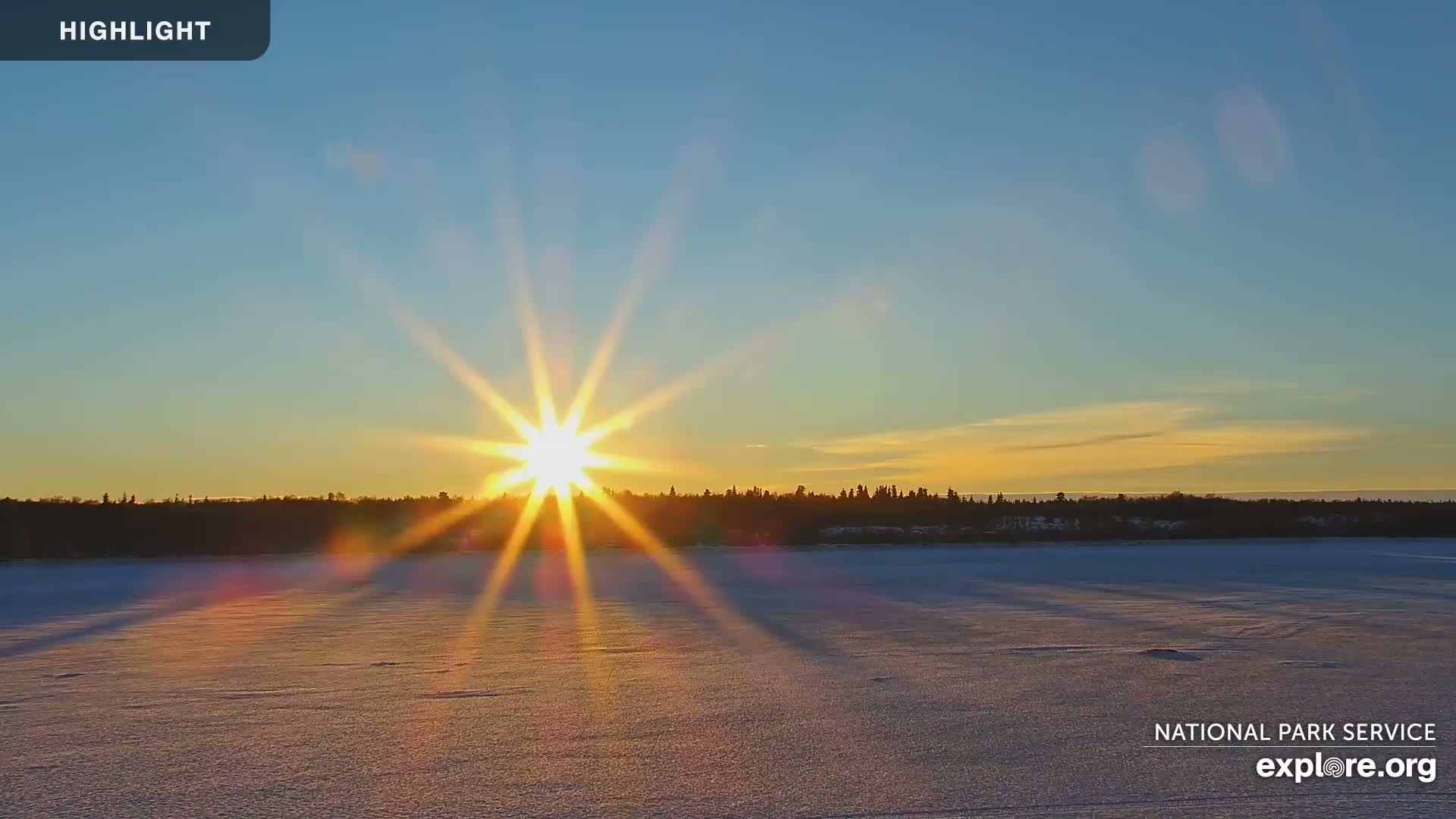A bright sun sets over a snow-covered frozen lake and a dark treeline, under a clear sky.