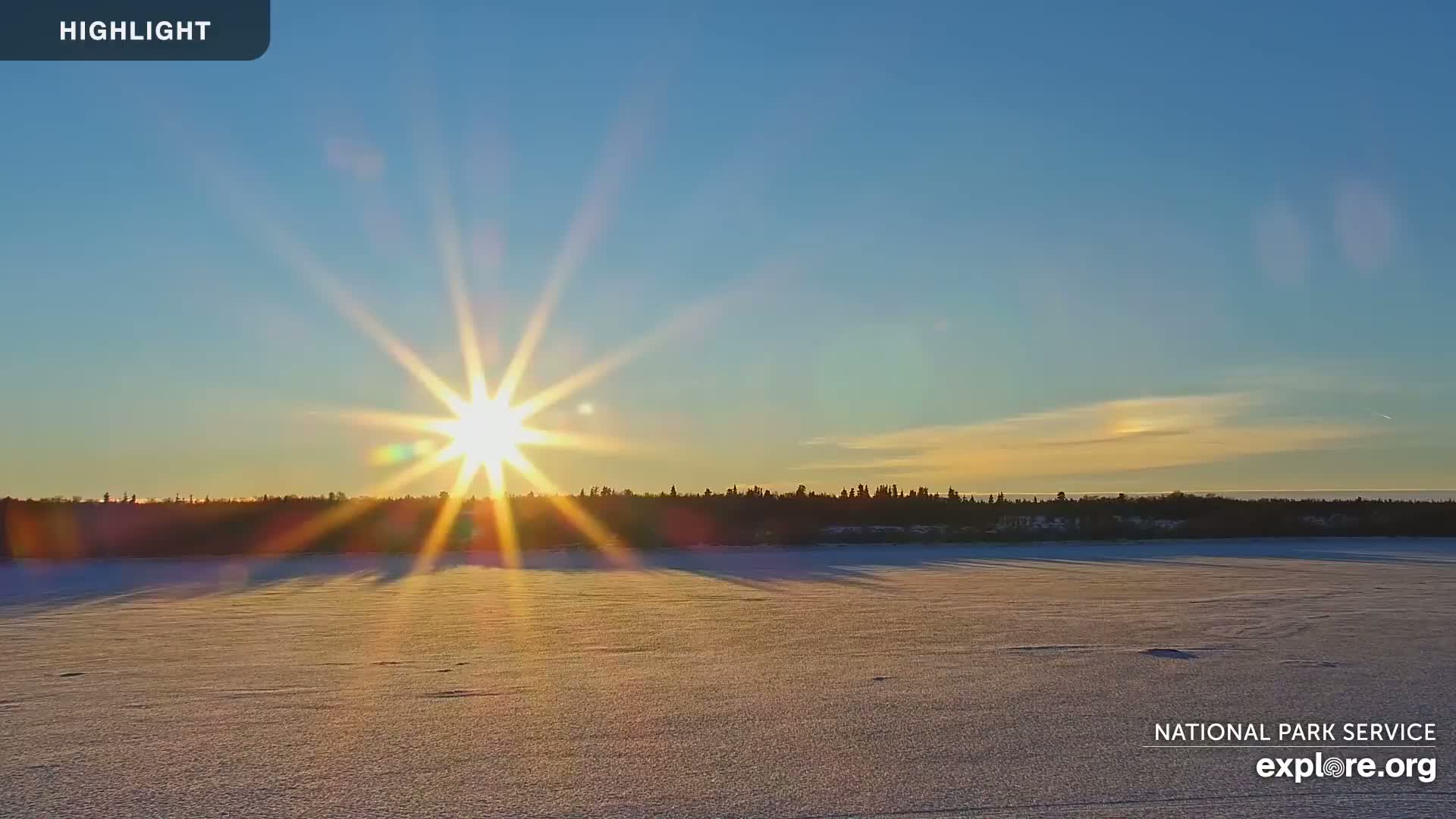A bright sun with lens flares sets over a snow-covered lake behind a dark treeline under a clear blue sky.