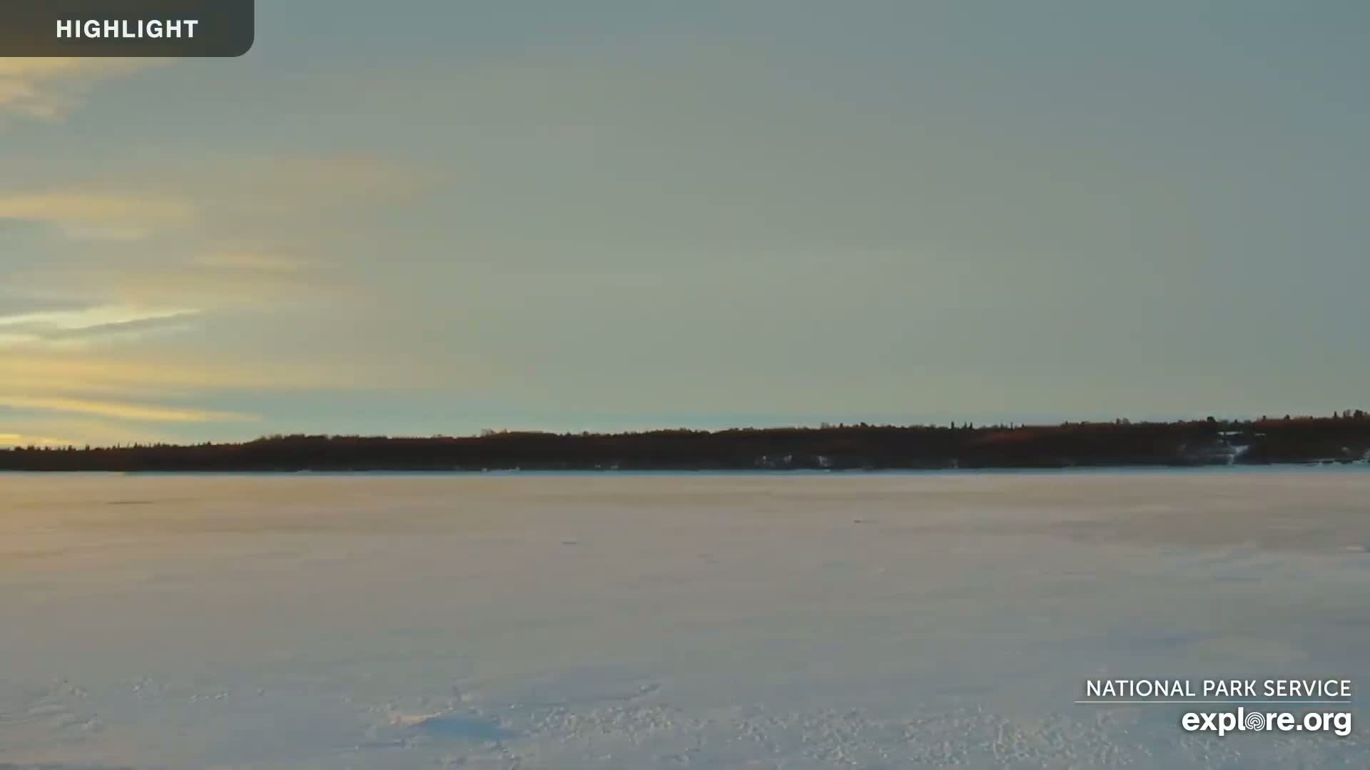 A frozen lake stretches to a treeline under a pale, partly cloudy sky.