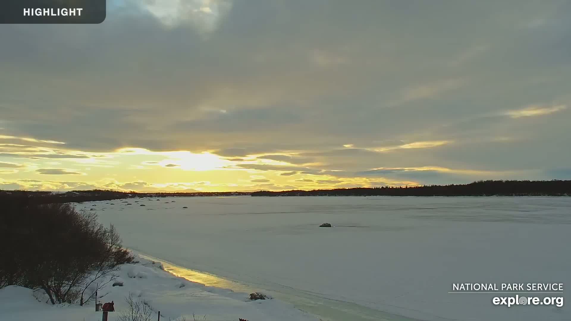 A frozen lake stretches to a distant treeline under a partly cloudy sky with a setting sun.
