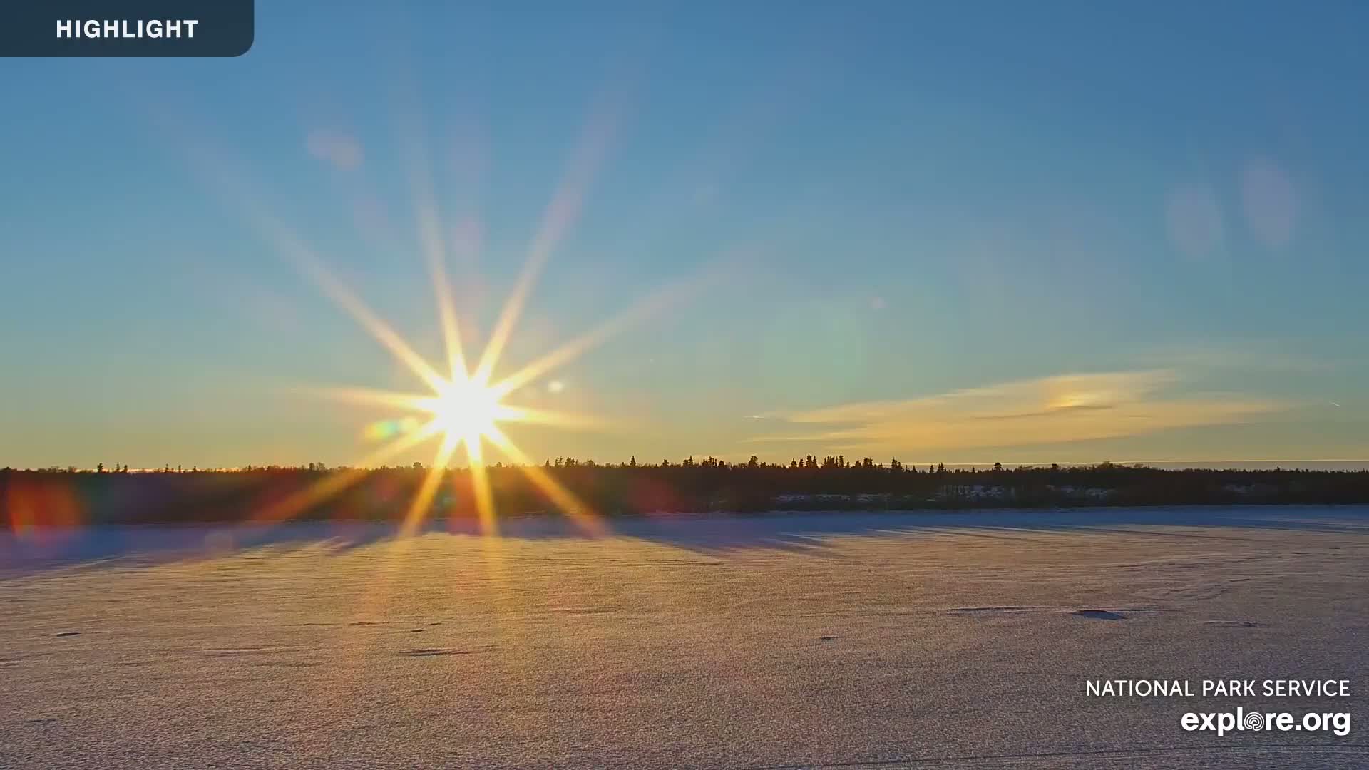 A bright sun shines over a frozen, snow-covered lake with a dark tree line on the horizon under a clear, mostly blue sky.