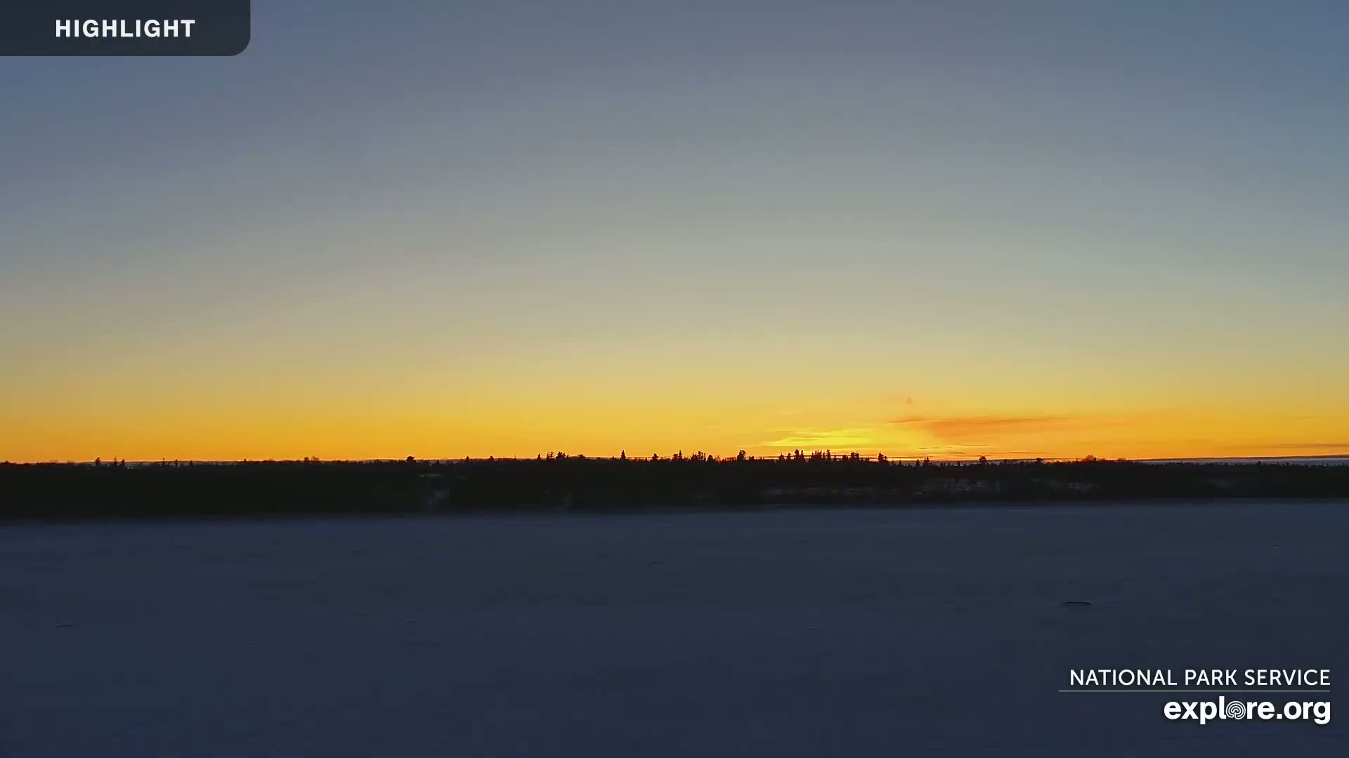 A snow-covered expanse stretches out before a dark treeline silhouetted against a vibrant orange and yellow sunset.