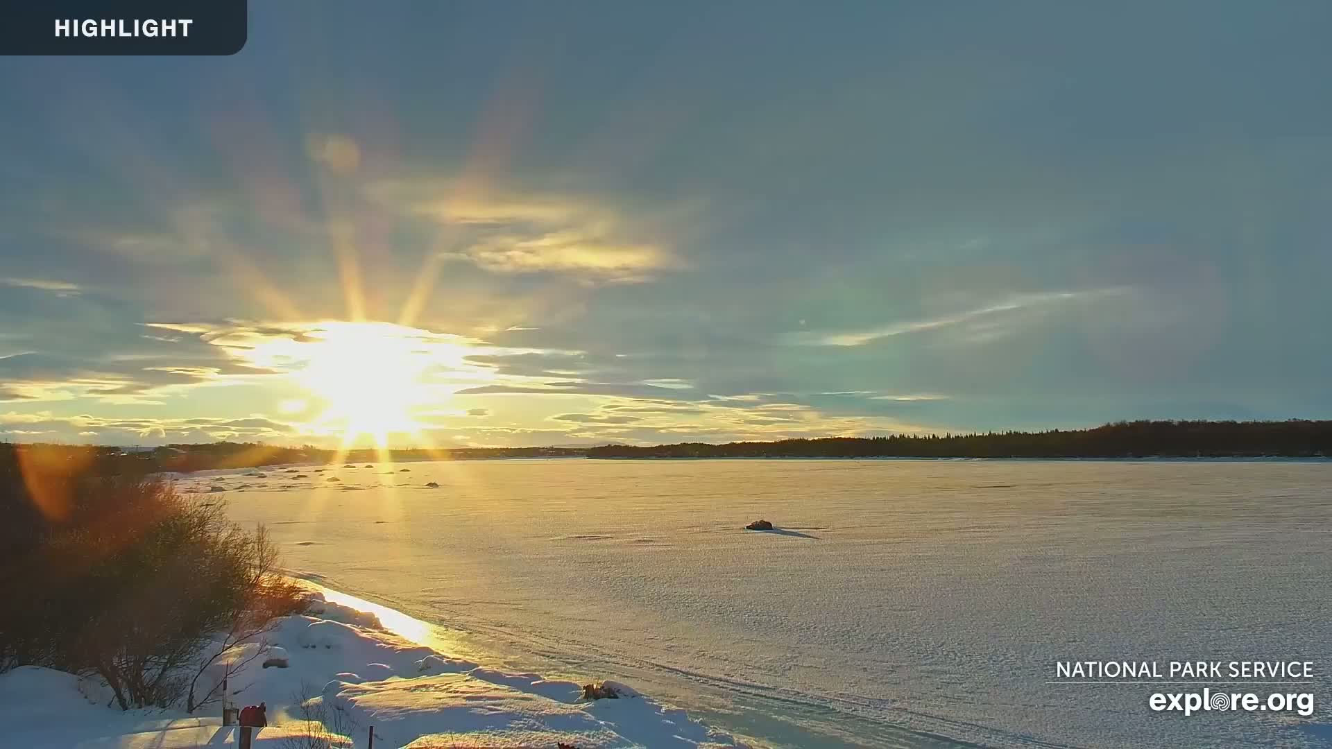 A bright, low sun sets over a frozen lake, casting a golden glow on the snow-covered landscape.
