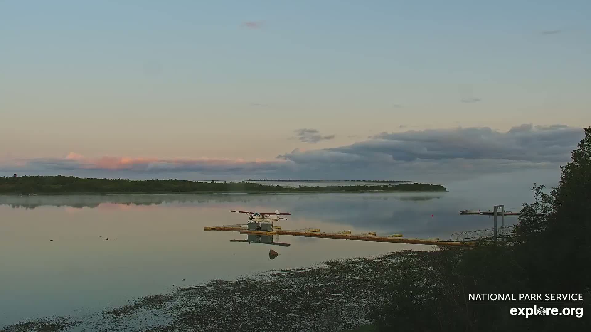 A seaplane sits on a dock in a calm lake at dawn, with low-lying fog over the water and a partly cloudy sky.