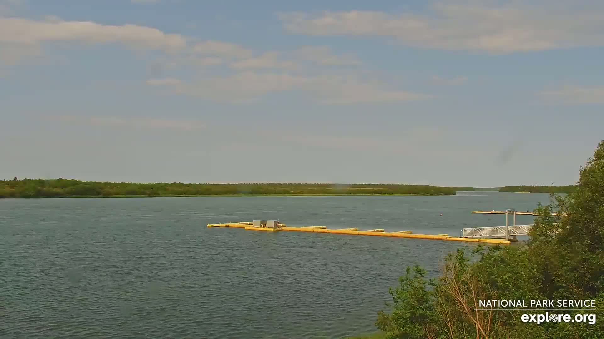 A calm river with a long, yellow floating dock extends from a small, metal pier under a partly cloudy sky.