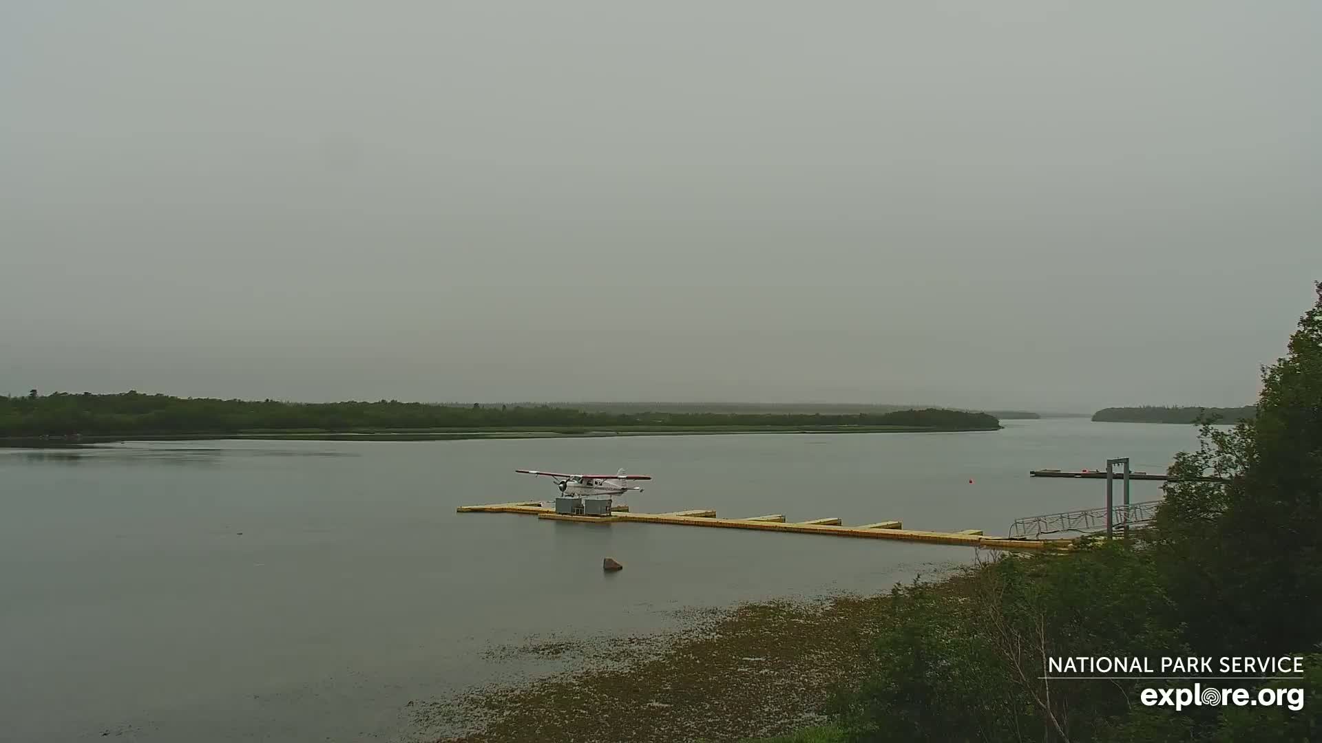 A seaplane sits on a dock in a calm river, under an overcast sky.