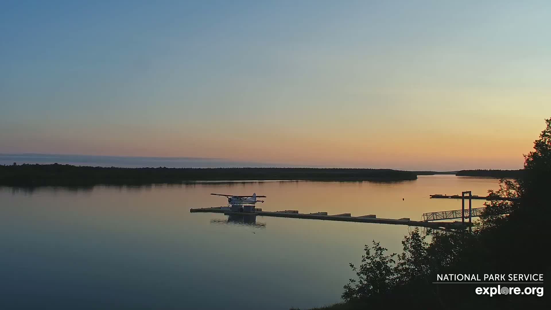 A seaplane rests on a dock on a calm lake at sunset, with a tree line visible in the distance.