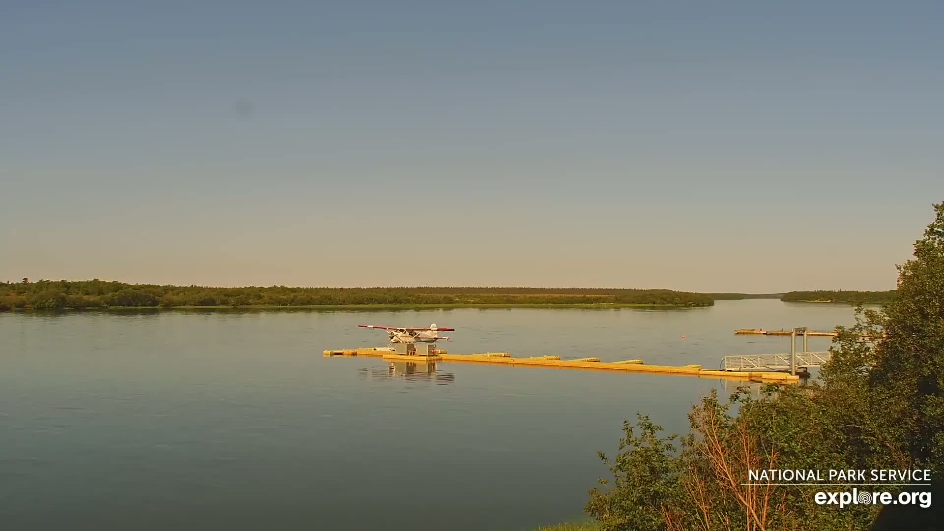 A seaplane is docked at a floating platform on a calm river under a clear, light-blue sky.