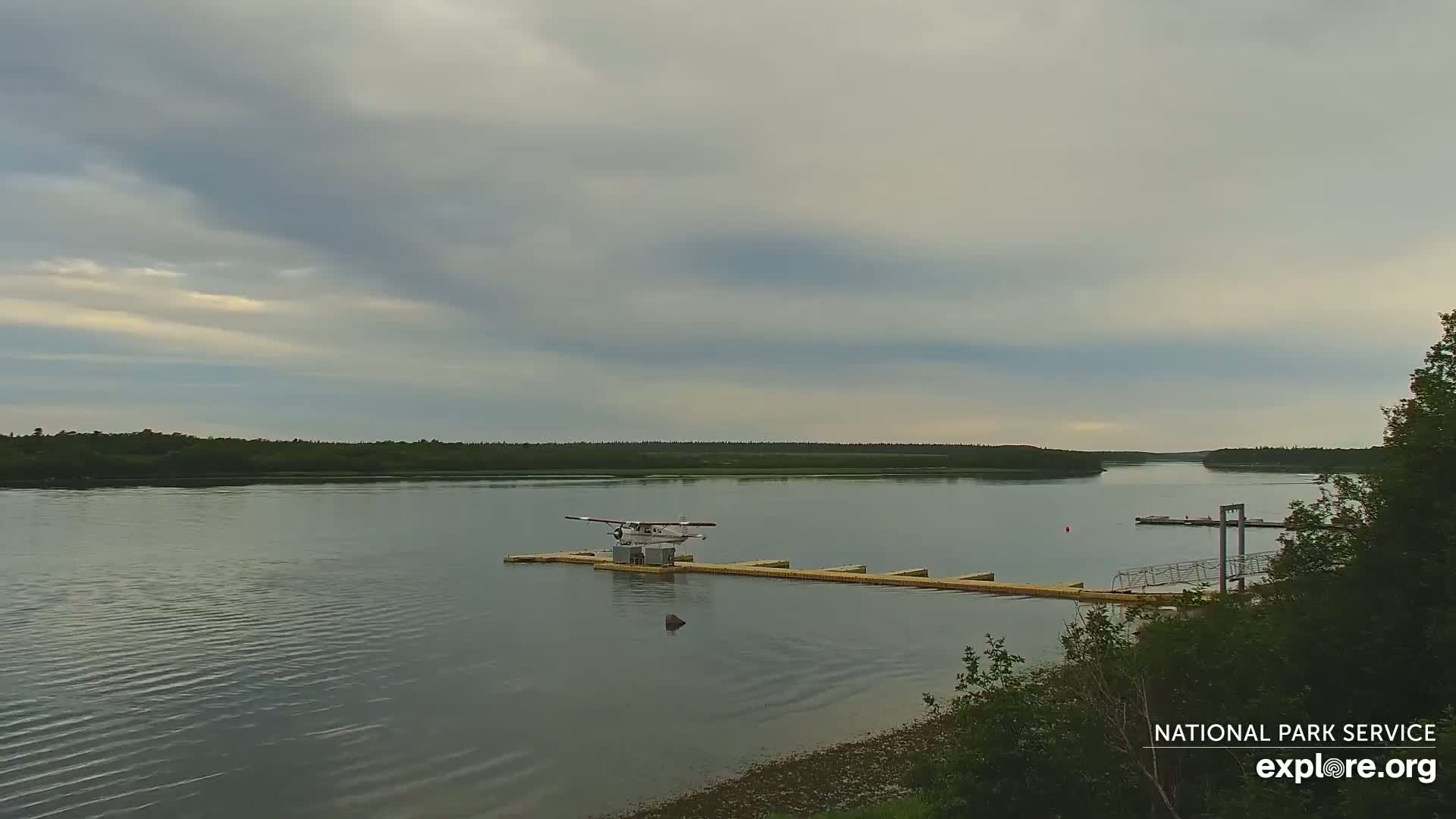 A seaplane sits on a dock on a calm river under a mostly cloudy sky.