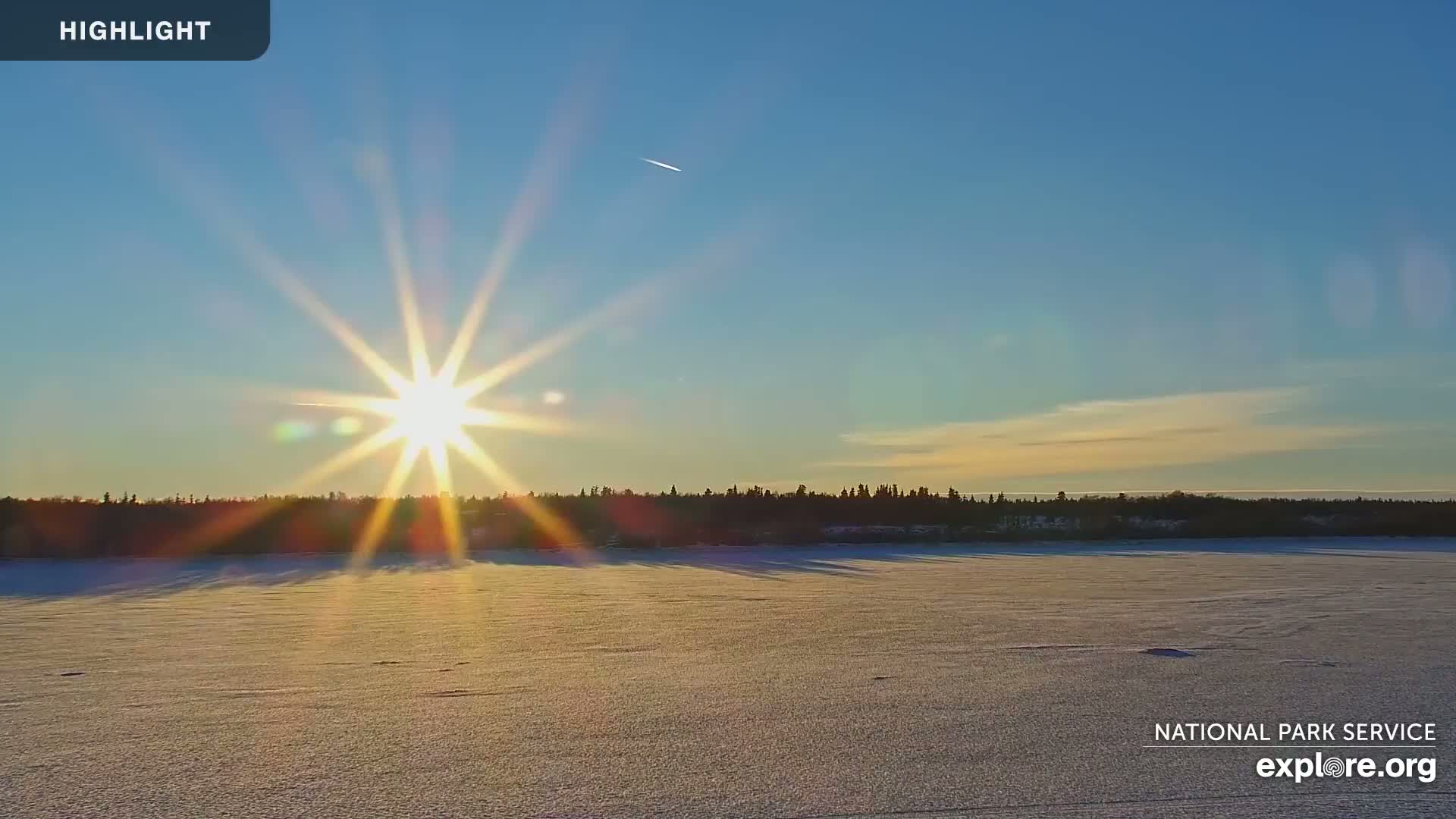 A bright sun shines over a frozen lake with a dark treeline in the background under a clear blue sky.