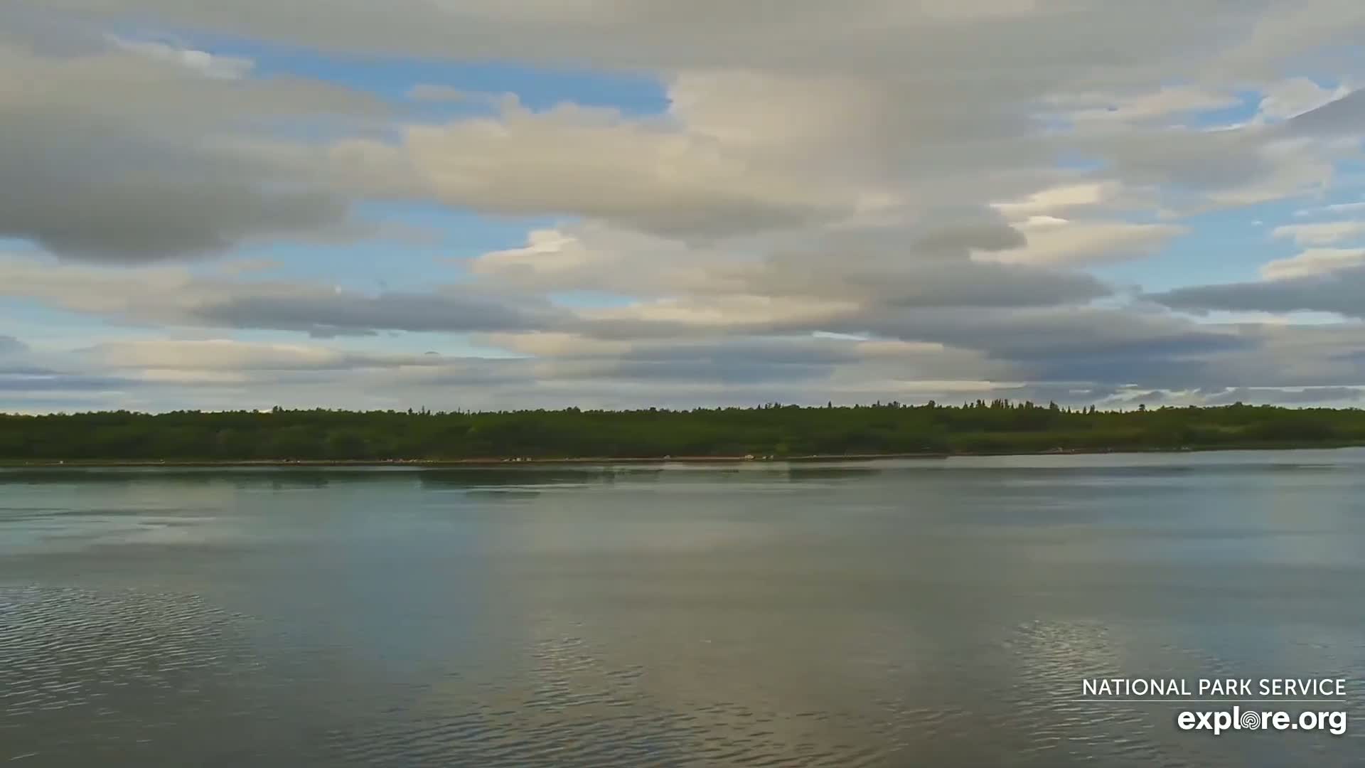 A calm body of water stretches across the foreground, reflecting a cloudy sky above a line of dark green trees in the distance.