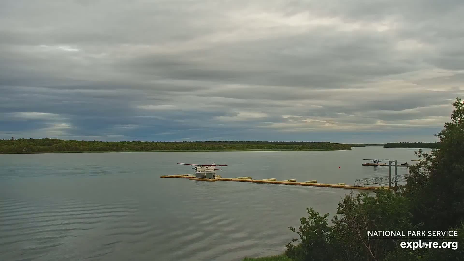 A seaplane sits on a dock in a calm lake under a cloudy sky, with another seaplane visible in the distance.
