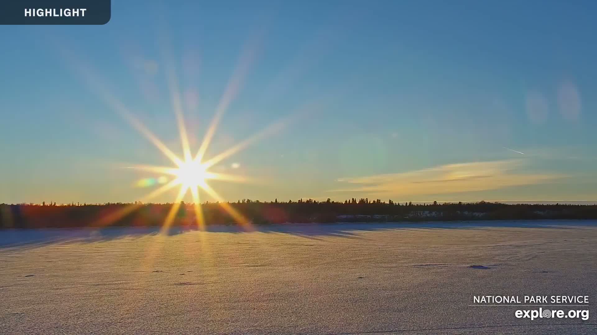 A brilliant sun low in the sky casts long shadows across a vast, snow-covered plain or frozen lake, bordered by a distant dark tree line under a clear blue sky with sparse clouds.
