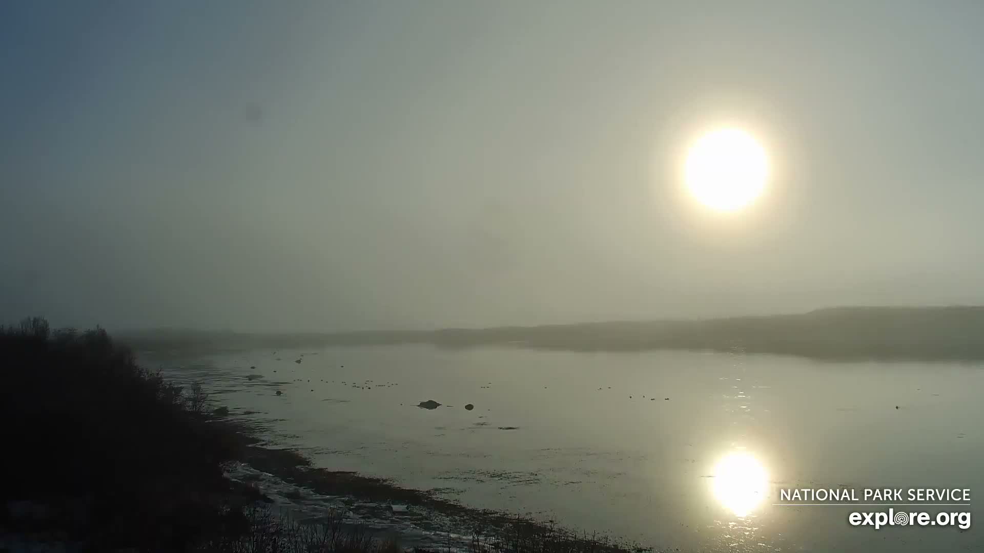 A hazy winter day shows a bright low sun reflecting on a wide body of water, with a dark, snow-dusted bank on the left and a distant hazy shore under a muted sky.