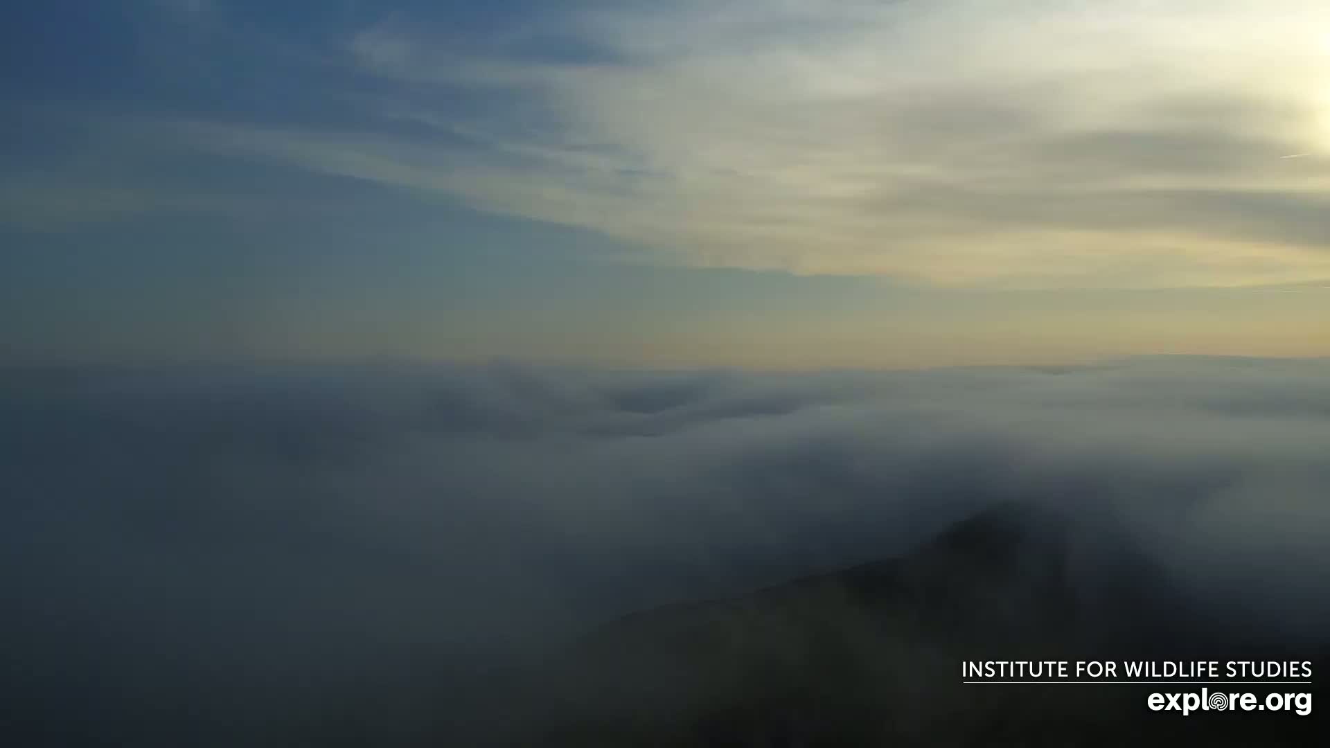 A high-altitude view shows a landscape shrouded in fog under a partly cloudy sky.
