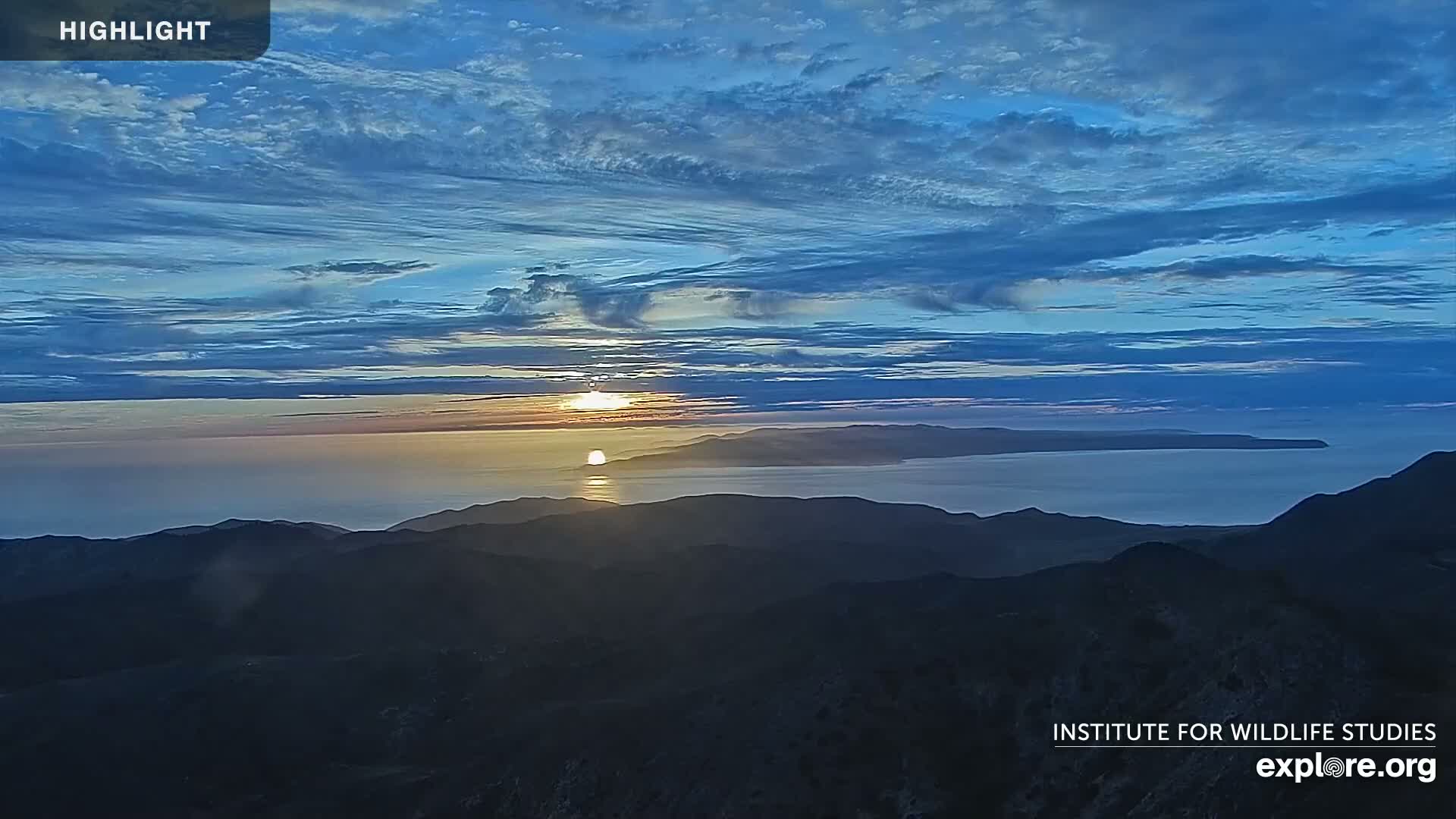 A sunrise over a calm ocean, viewed from a mountaintop under a mostly clear, blue sky with thin clouds.