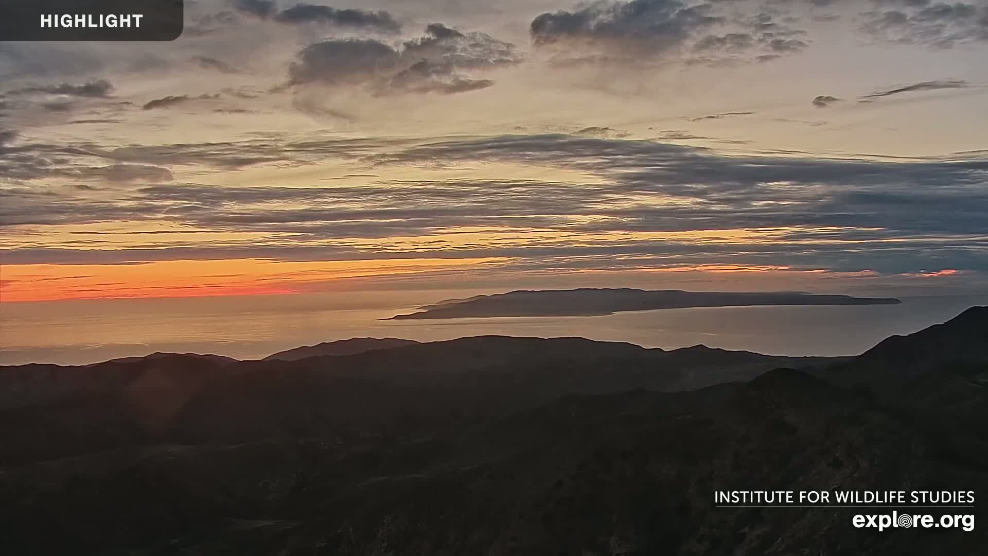 A sunset over a calm ocean and distant islands, viewed from a mountain range under a mostly cloudy sky.