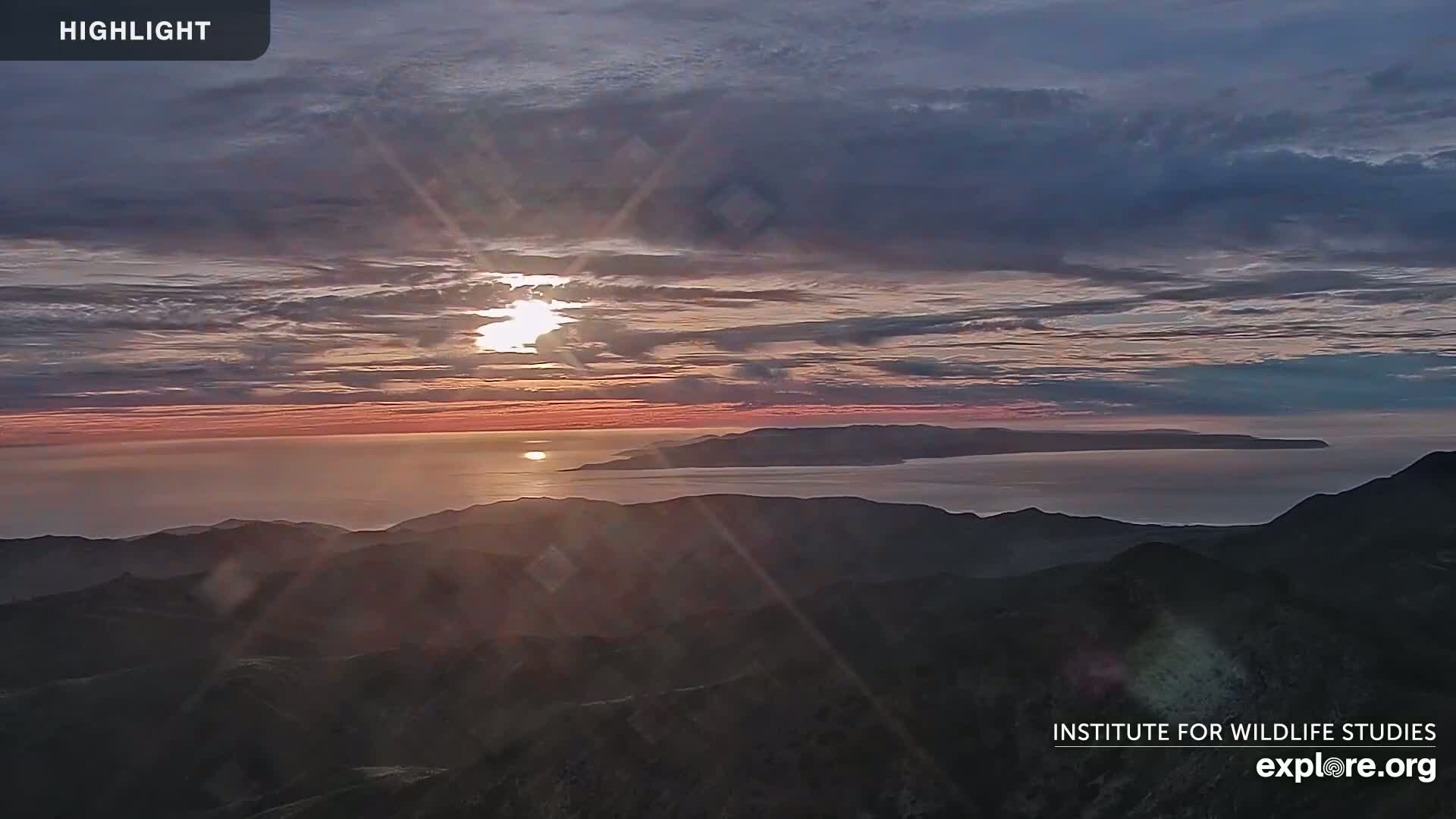 A sunrise over a calm ocean and distant island, viewed from a mountaintop under a partly cloudy sky.