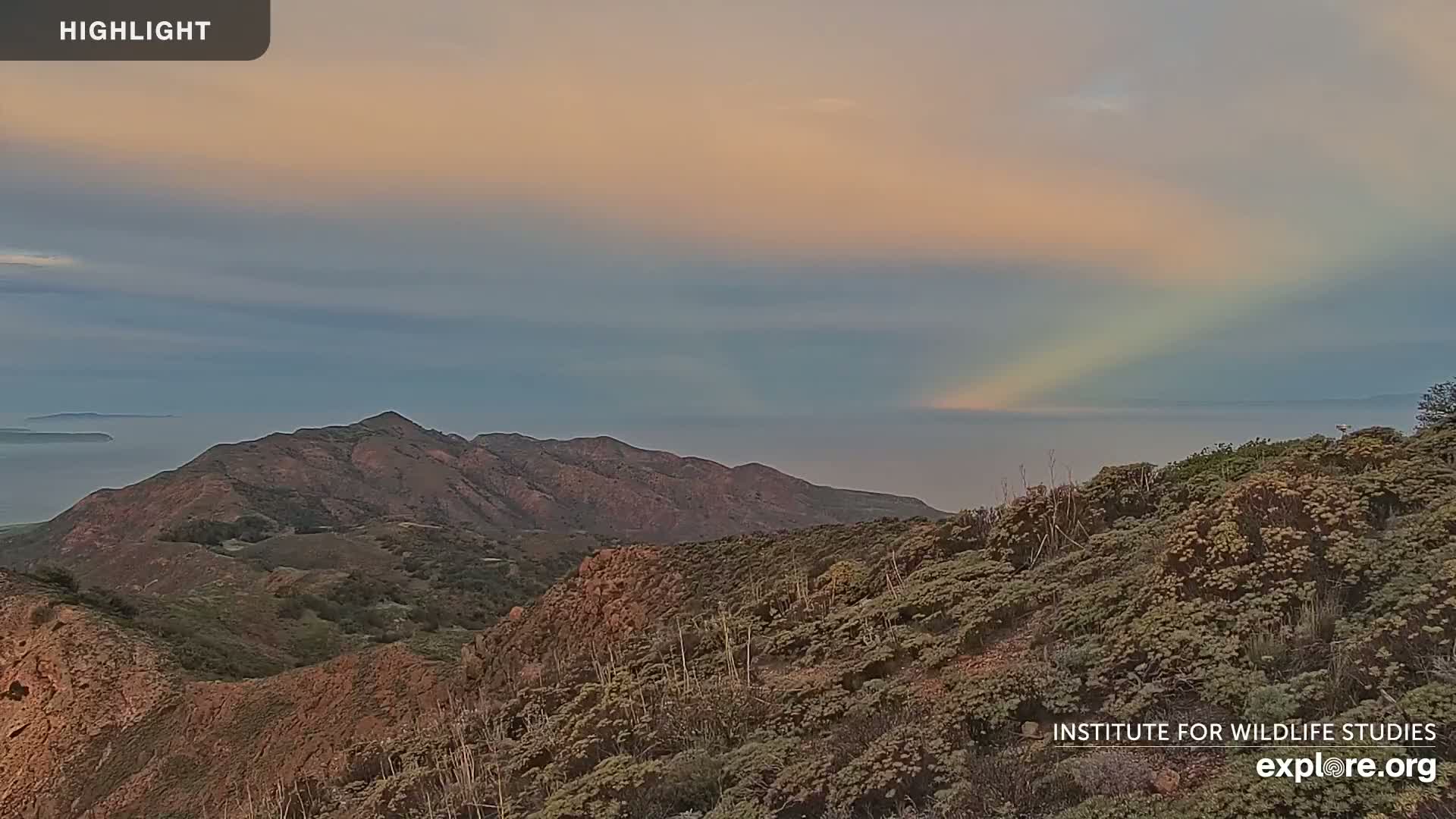 A faint rainbow appears in the sky over a mountainous landscape overlooking a calm ocean at sunset.