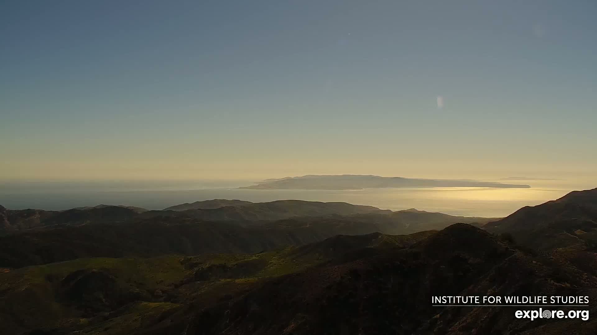 A tranquil sunset illuminates a calm ocean separating a distant island from a mountainous foreground.