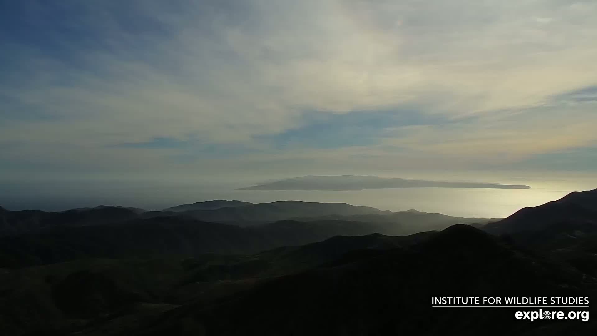 A hazy, distant island is visible across a calm ocean from a mountain range shrouded in shadow under a mostly cloudy sky.