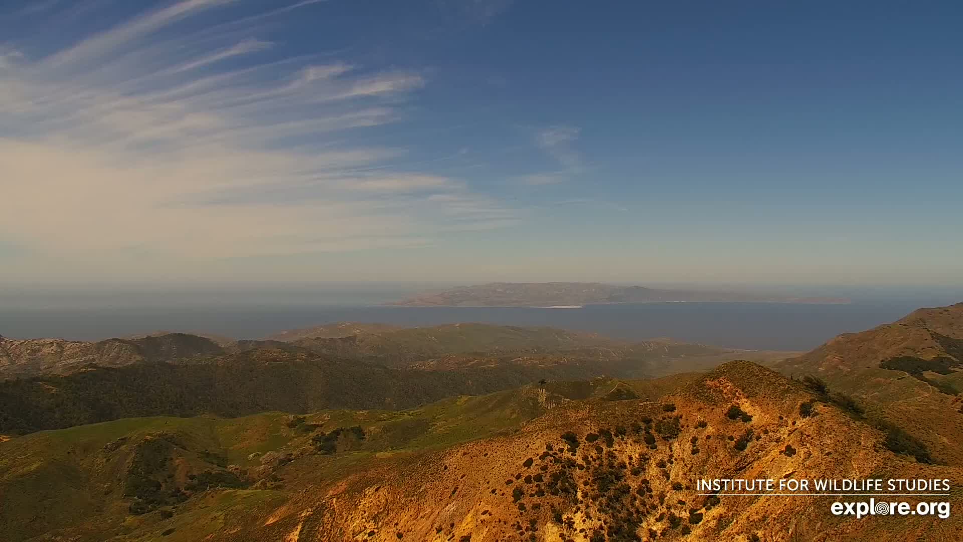 A high-angle view shows rolling hills under a mostly clear, light-blue sky with thin, wispy clouds, overlooking a distant island and ocean.