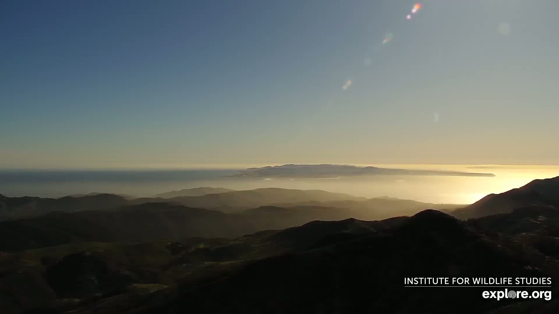 A hazy sunset illuminates layers of mountains and a distant island over a calm ocean.