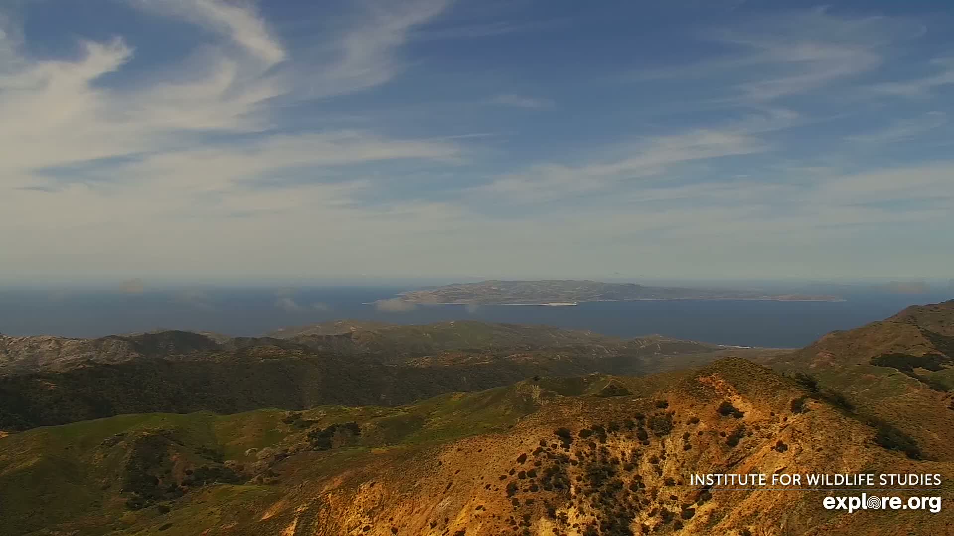 A high-angle view shows a mountainous landscape under a mostly clear sky with scattered clouds, overlooking a calm ocean with an island in the distance.