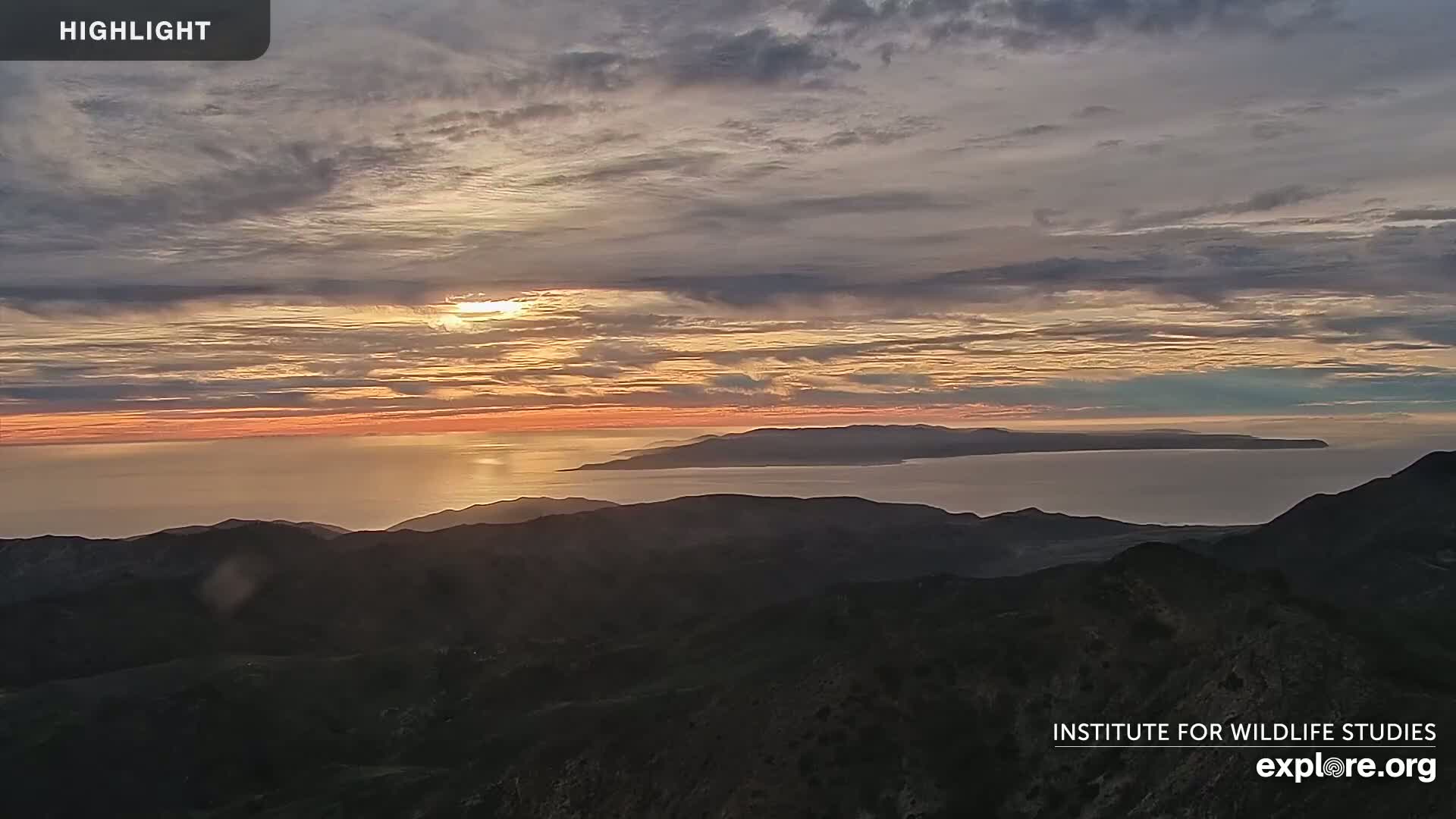 A sunset over a calm ocean and distant islands, viewed from a mountain range under a cloudy sky.