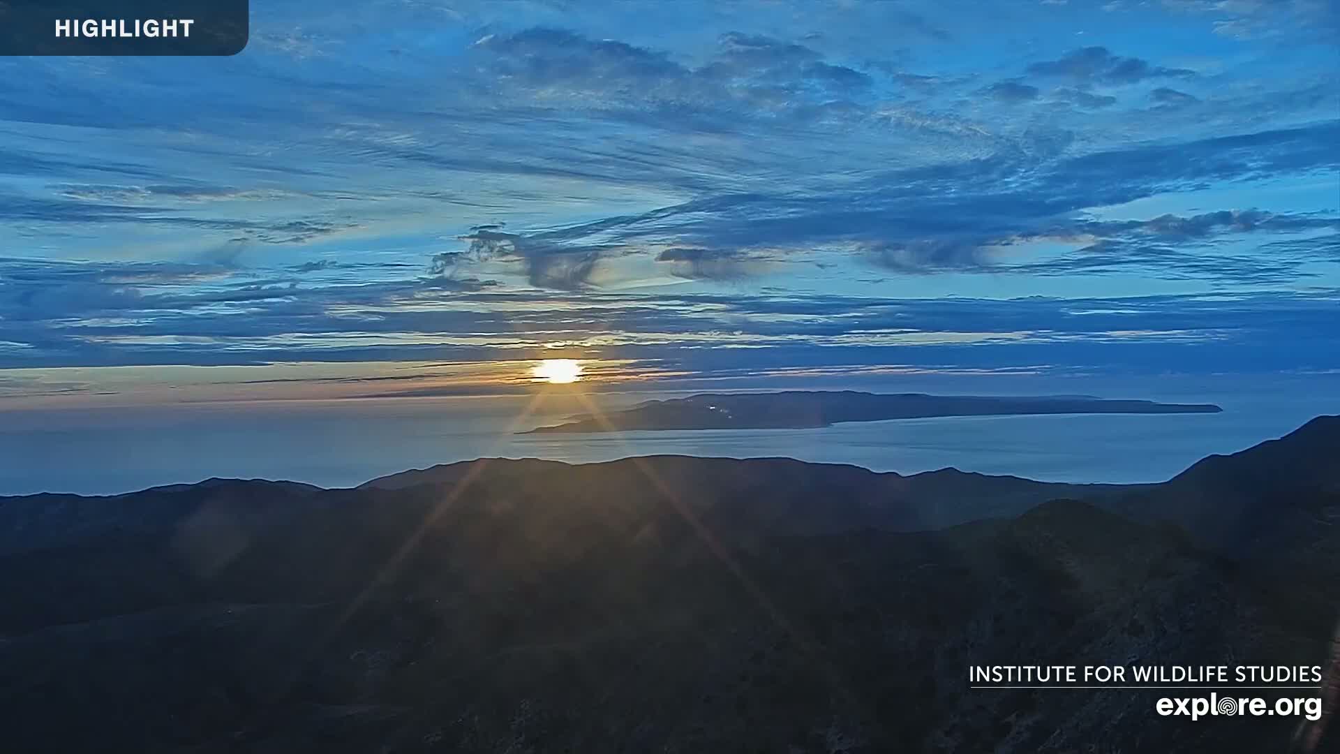 A sunrise over a calm ocean and distant islands, viewed from a mountaintop under a mostly clear, blue sky with thin clouds.