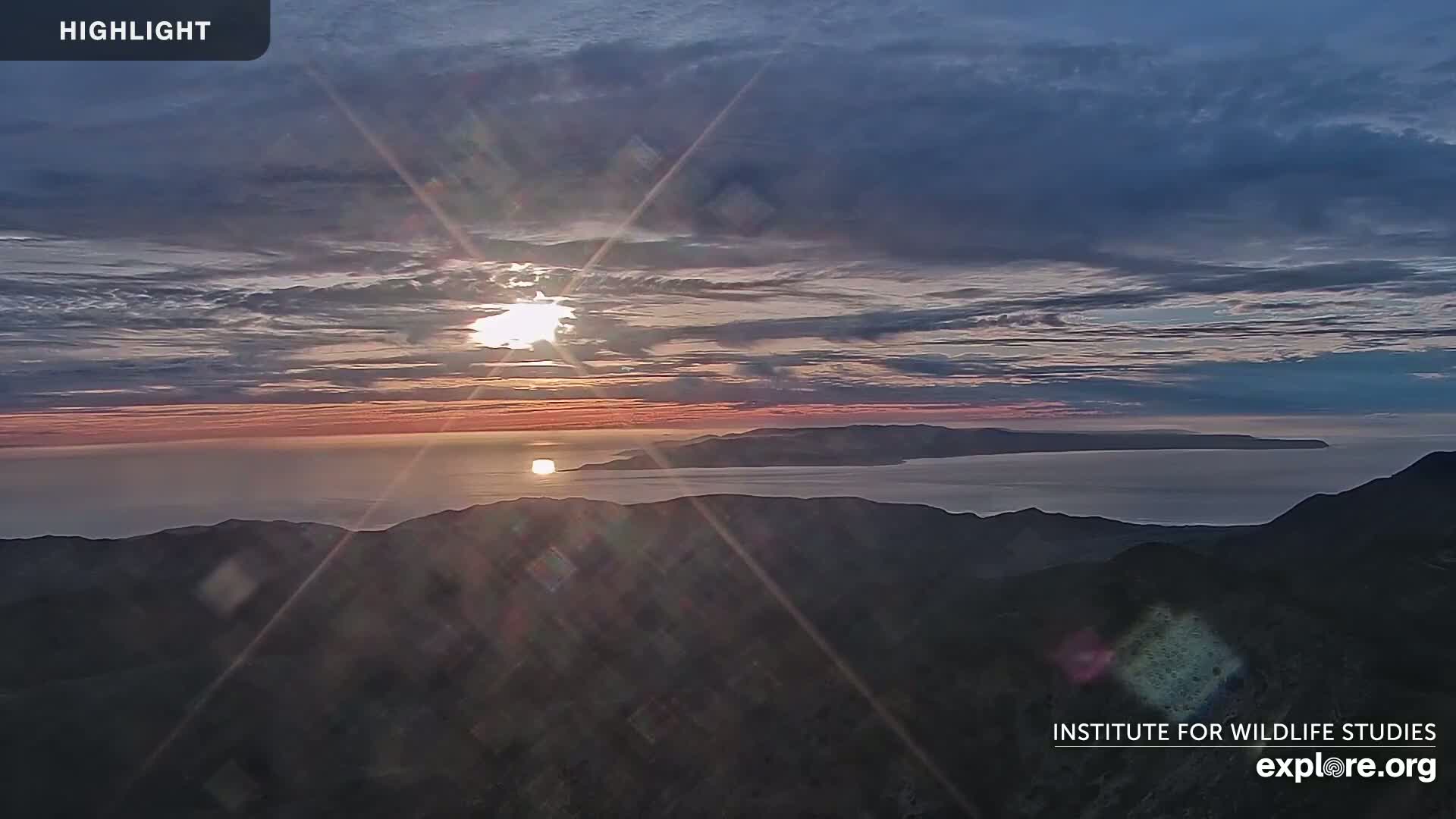 A sunrise over a calm ocean and distant islands is viewed from a mountaintop under a partly cloudy sky.