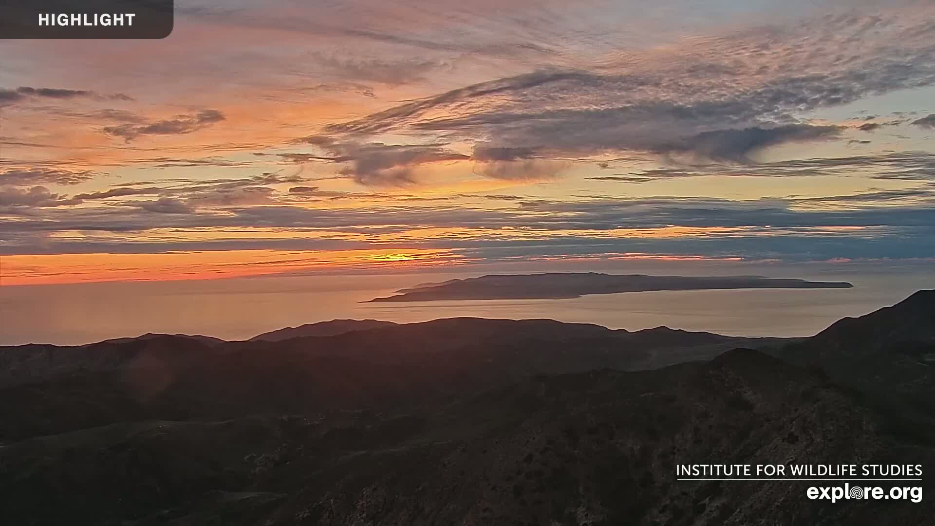 A sunset over a calm ocean and distant islands is viewed from a mountain range under a partly cloudy sky.