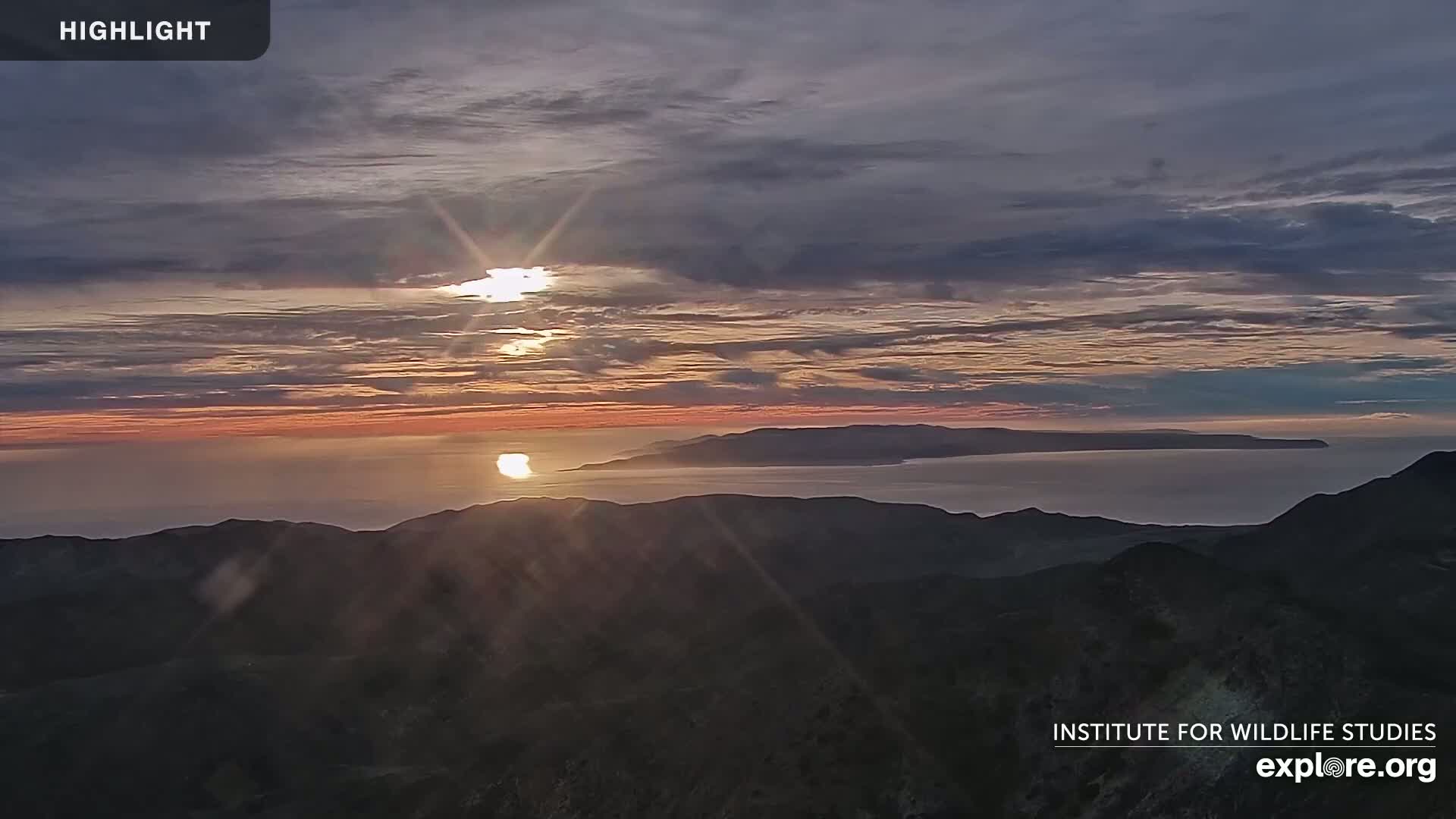 A sunrise over a calm ocean and distant island, viewed from a mountaintop under a partly cloudy sky.