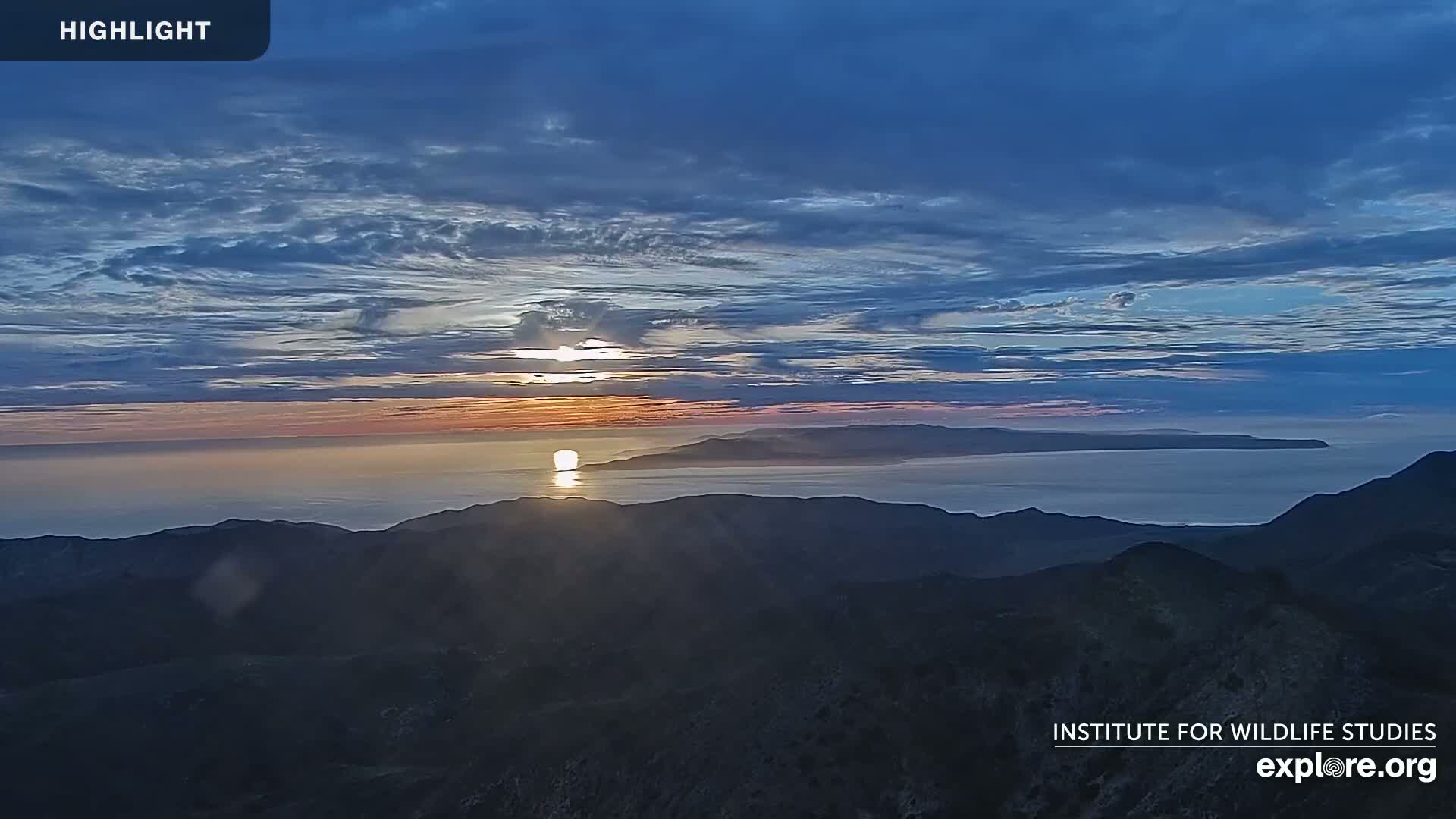 A sunset over a calm ocean, with a distant island visible, is seen from a mountaintop under a partly cloudy sky.
