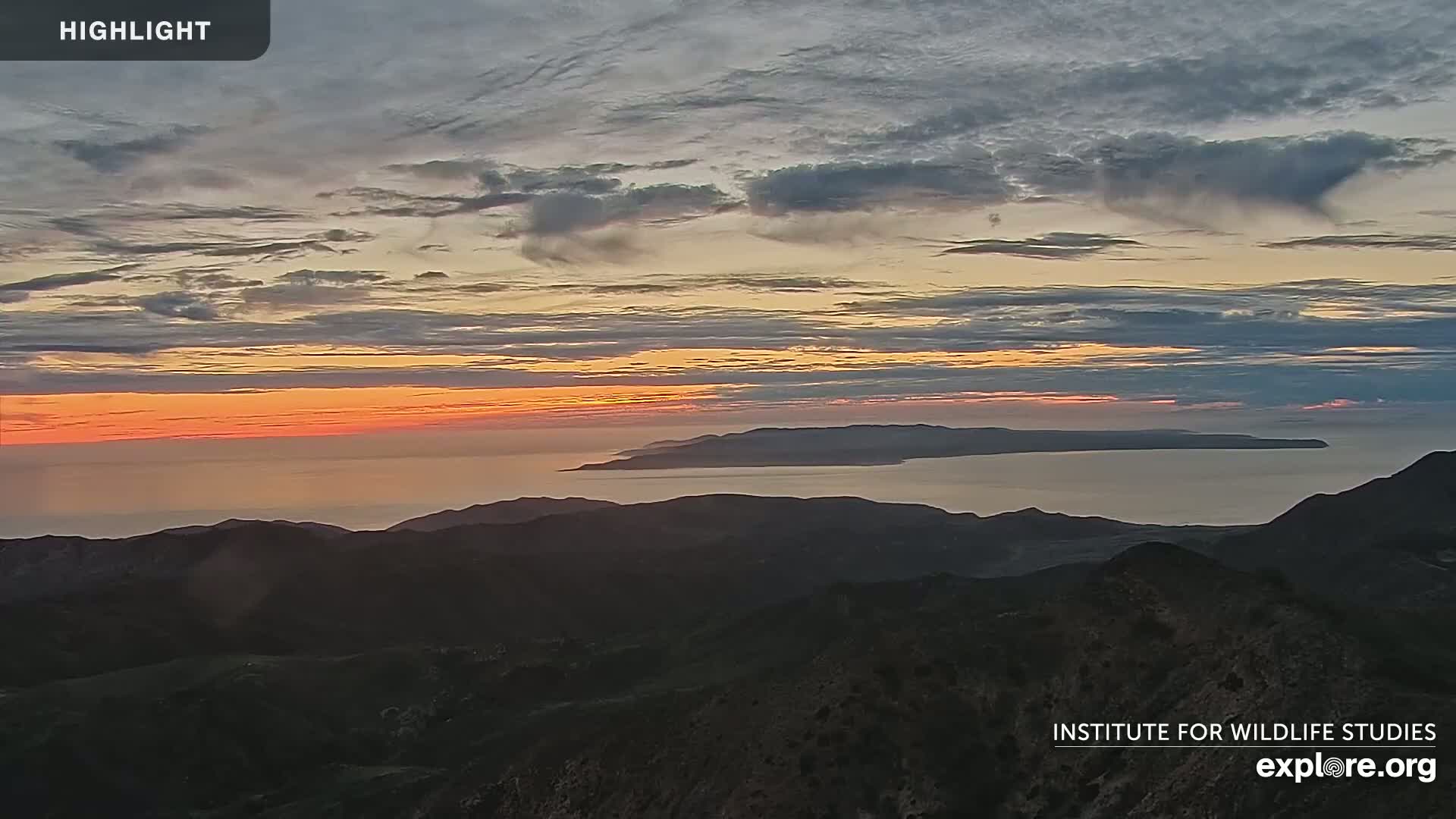 A sunset casts an orange glow over a calm ocean separating dark mountain ranges.