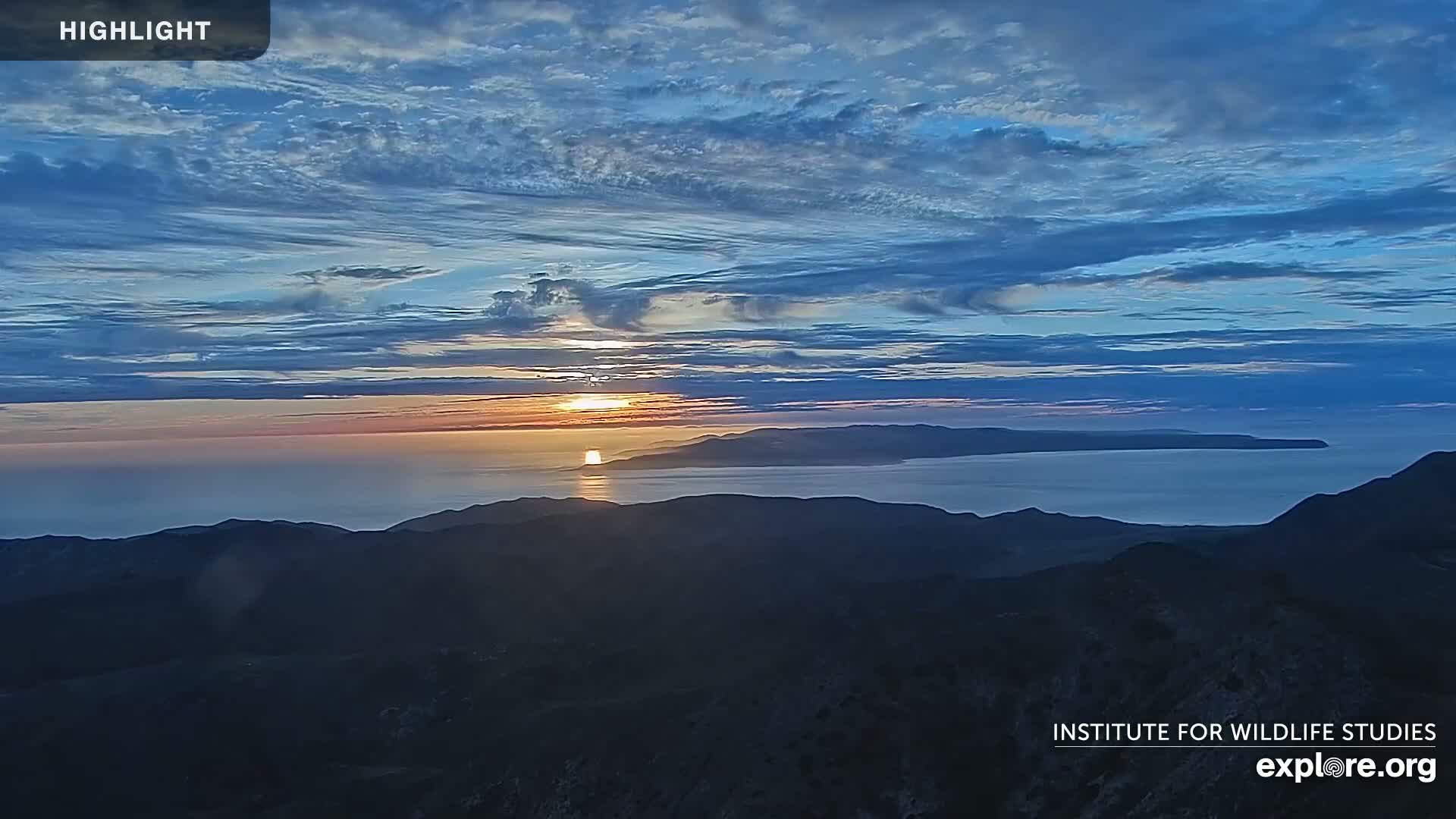 A sunrise over a calm ocean and distant islands is viewed from a mountaintop under a mostly clear sky with thin clouds.