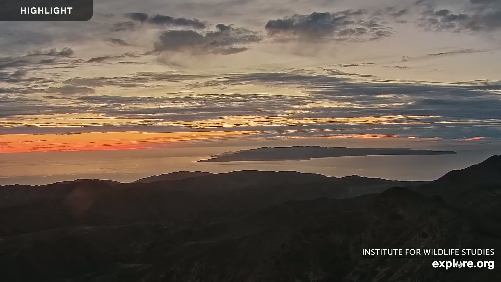 A hazy sunrise casts an orange glow on the ocean and islands, viewed from a mountain range.