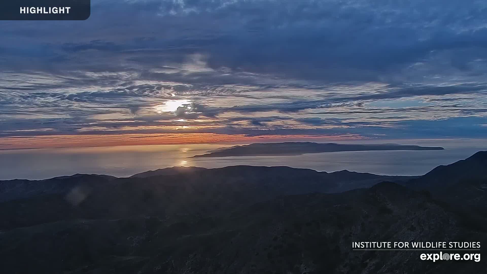 A sunrise over a calm ocean and distant islands, viewed from a mountaintop under a partly cloudy sky.
