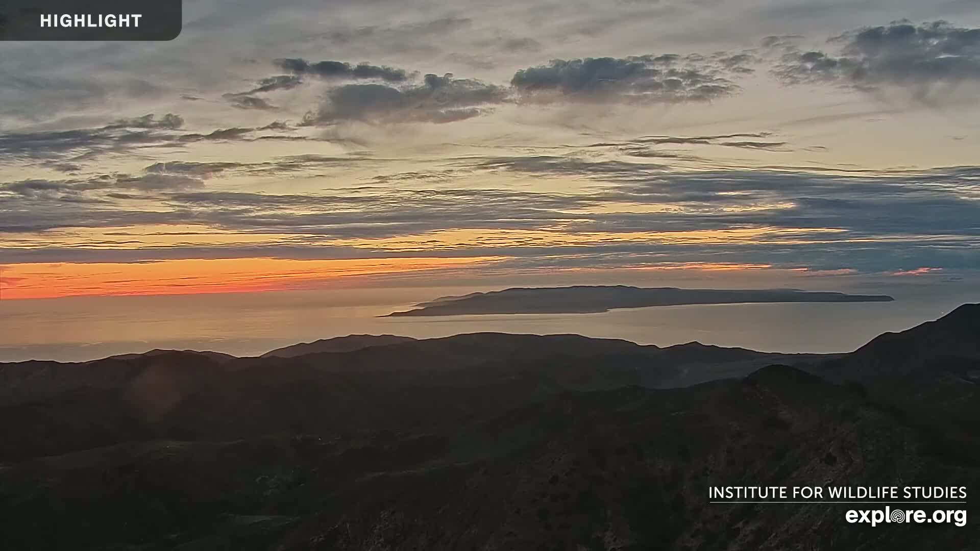 A sunset over a calm ocean with an island in the distance, viewed from a mountain range.