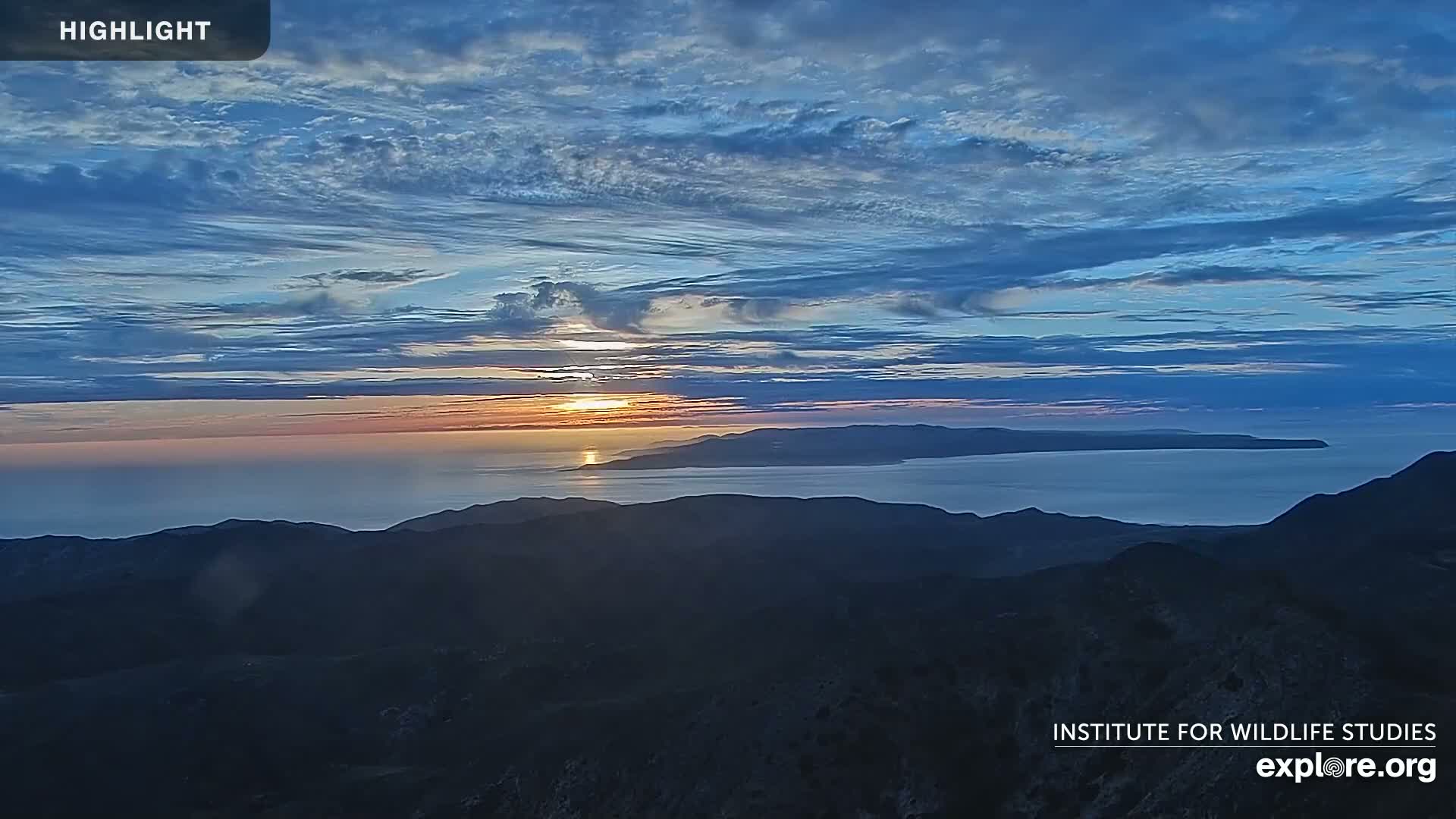 A sunrise over a calm ocean, with a distant island and mountainous foreground under a partly cloudy sky.