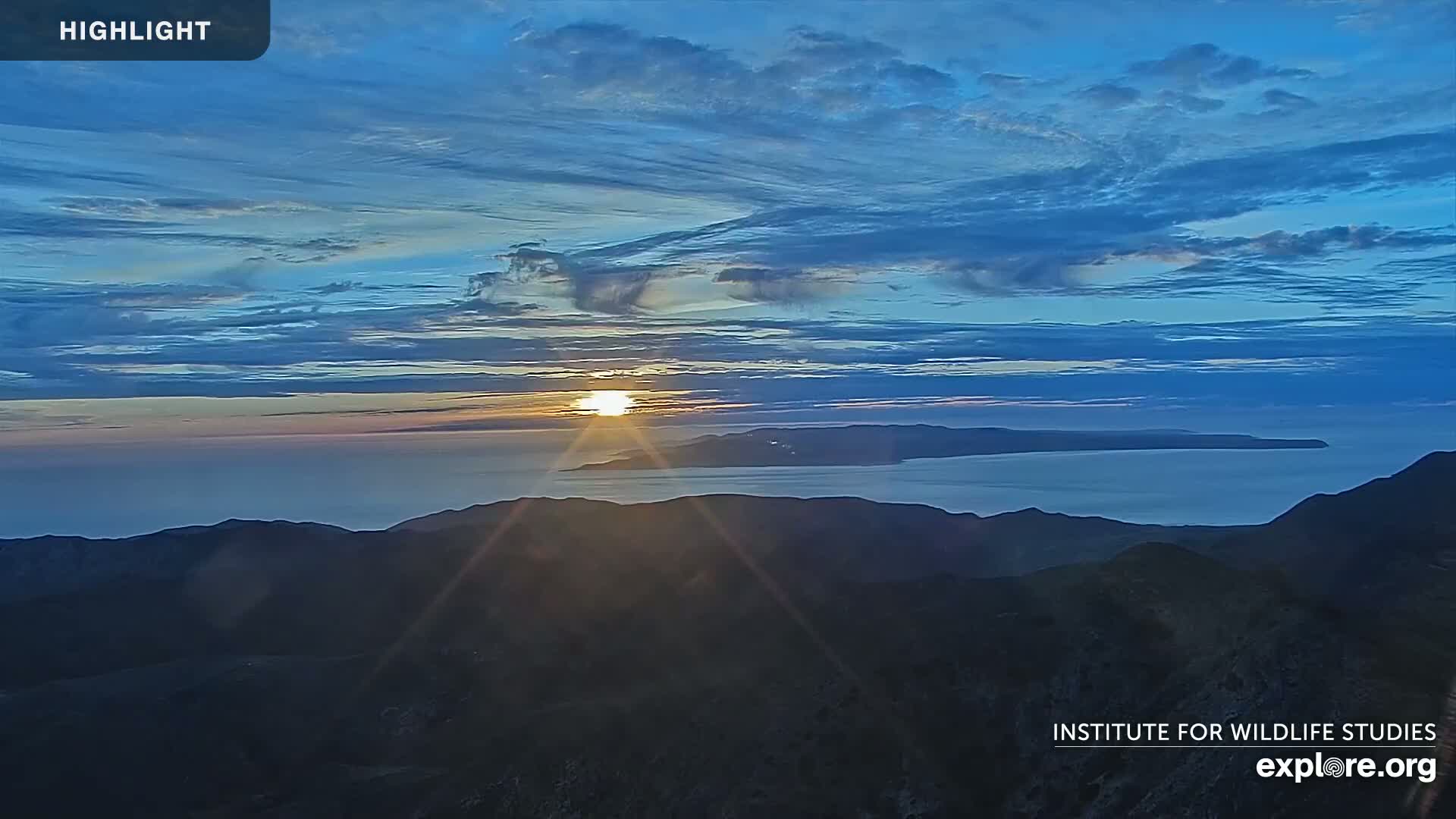 A sunrise over a calm ocean and distant islands, viewed from a mountaintop under a mostly clear, blue sky.