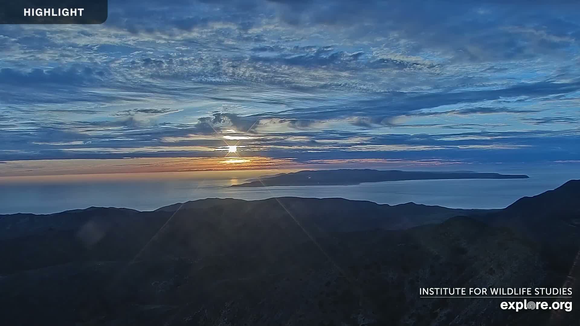 A sunset over a calm ocean and island, viewed from a mountaintop under a mostly clear, blue sky with high clouds.