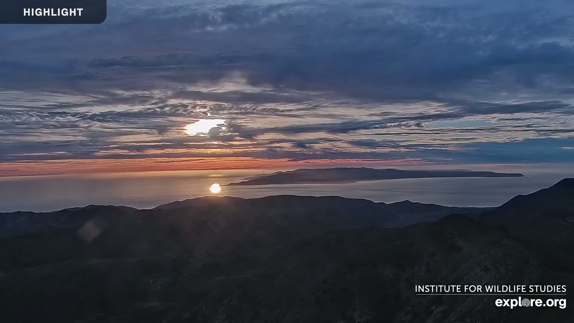 A sunset over a calm ocean and distant islands, viewed from a mountaintop under a mostly cloudy sky.