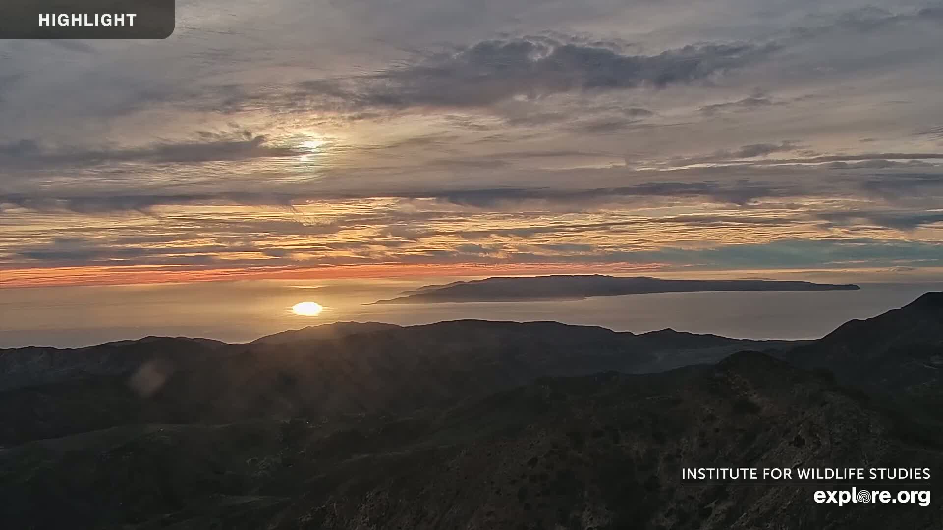 A sunset over a calm ocean, viewed from a mountain range, with a partly cloudy sky showing streaks of orange and pink.