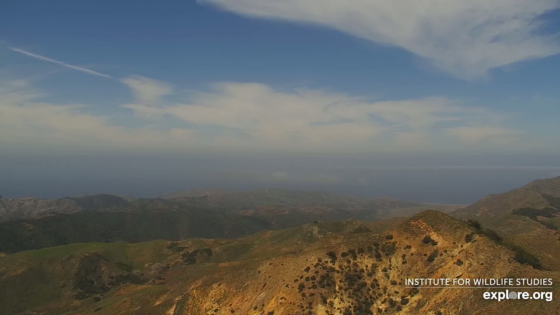 A high-angle view shows a mostly sunny day over a range of brown and green hills overlooking a hazy ocean.