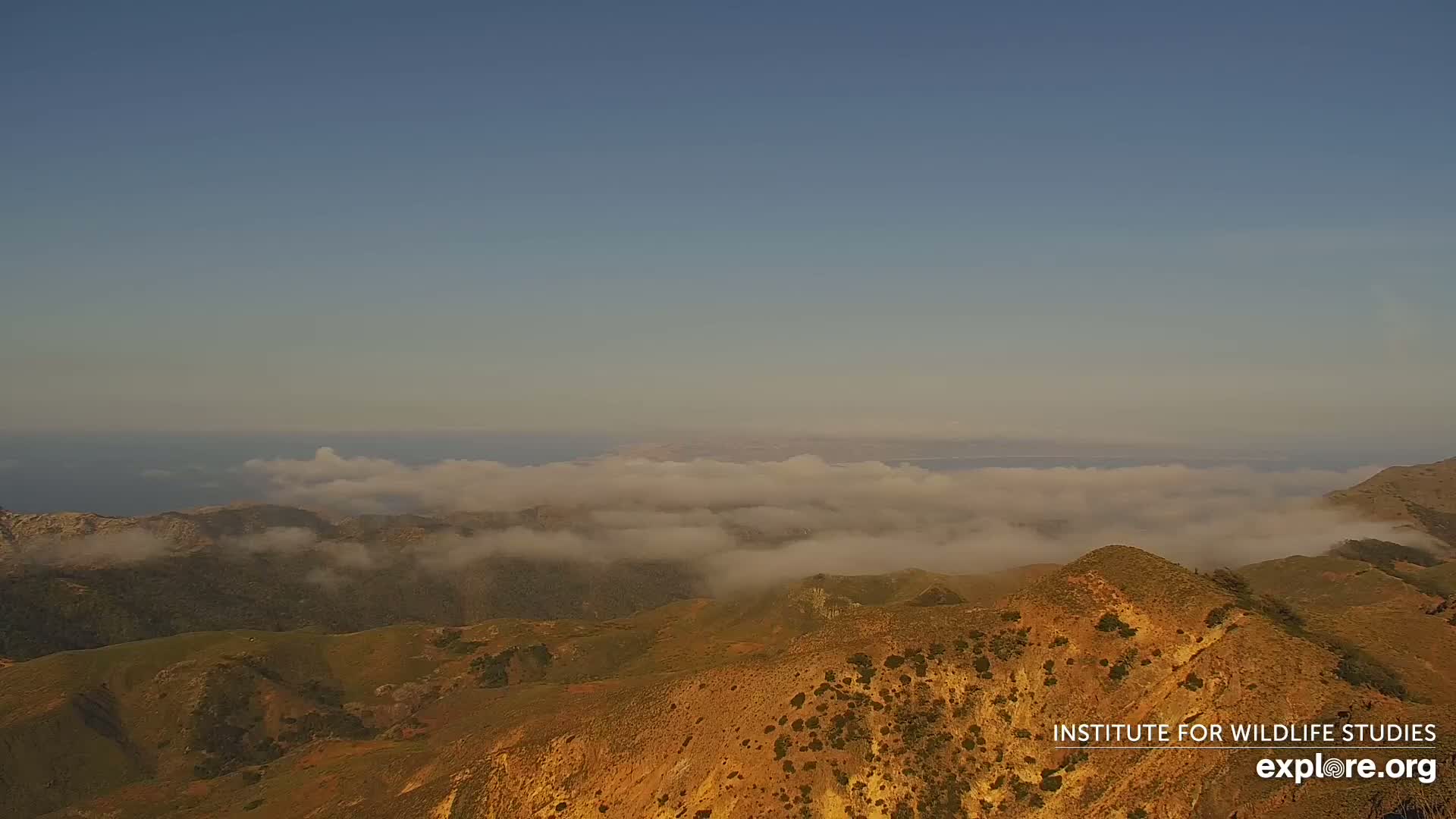 A high-altitude view shows rolling, tan-colored hills partially obscured by low-lying clouds under a clear, pale blue sky, with a body of water visible in the distance.
