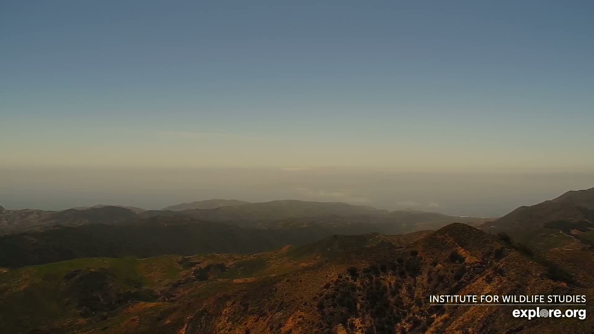 A hazy, distant ocean is visible beyond a mountainous landscape of brown and green hills.