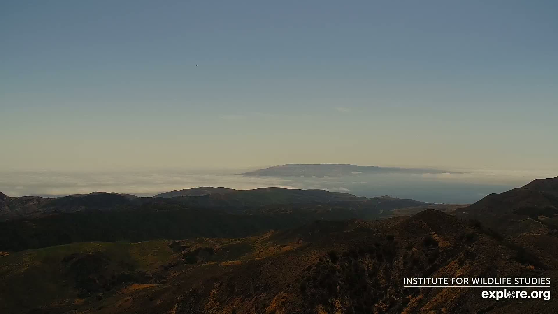 A high-angle view shows rolling hills and mountains shrouded in low-lying clouds, with a distant island visible through the hazy atmosphere.