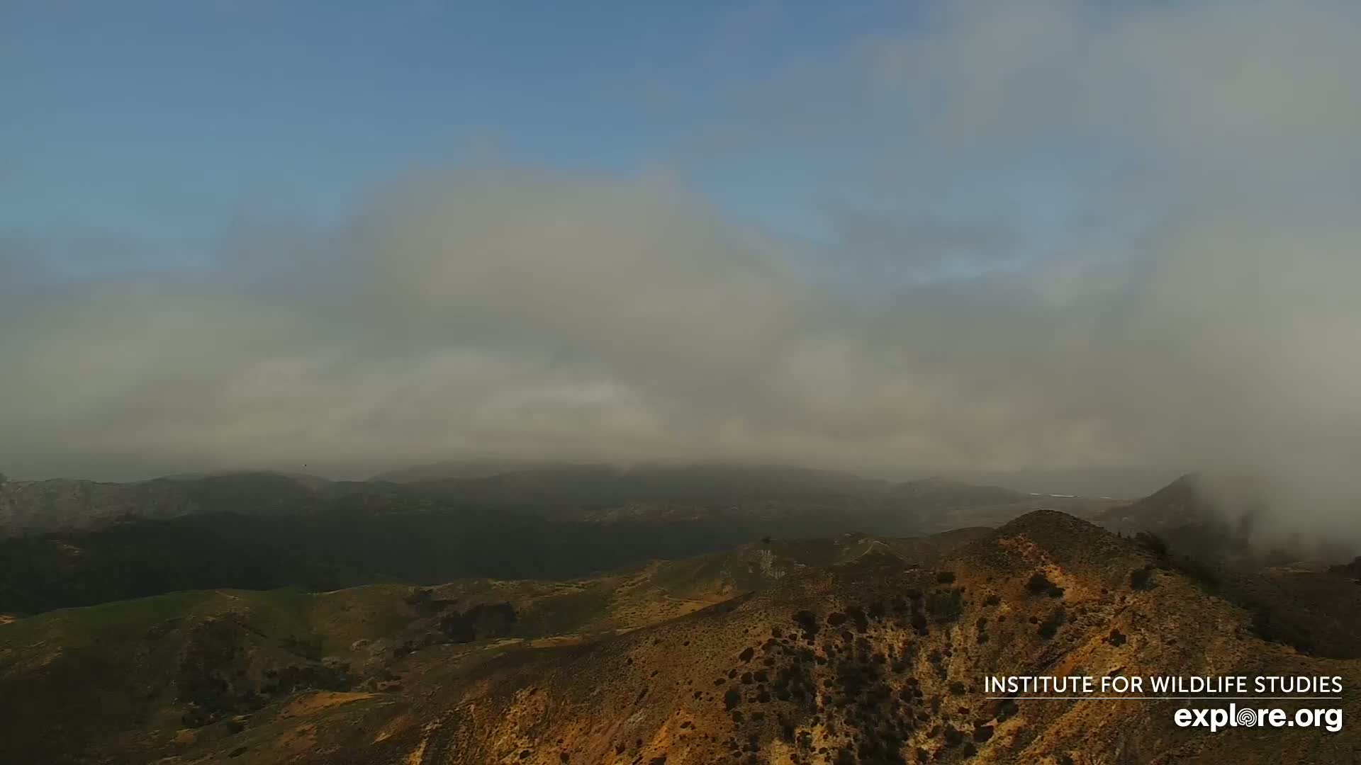 A high-angle view shows rolling hills and mountains partially obscured by fog under a mostly clear sky.
